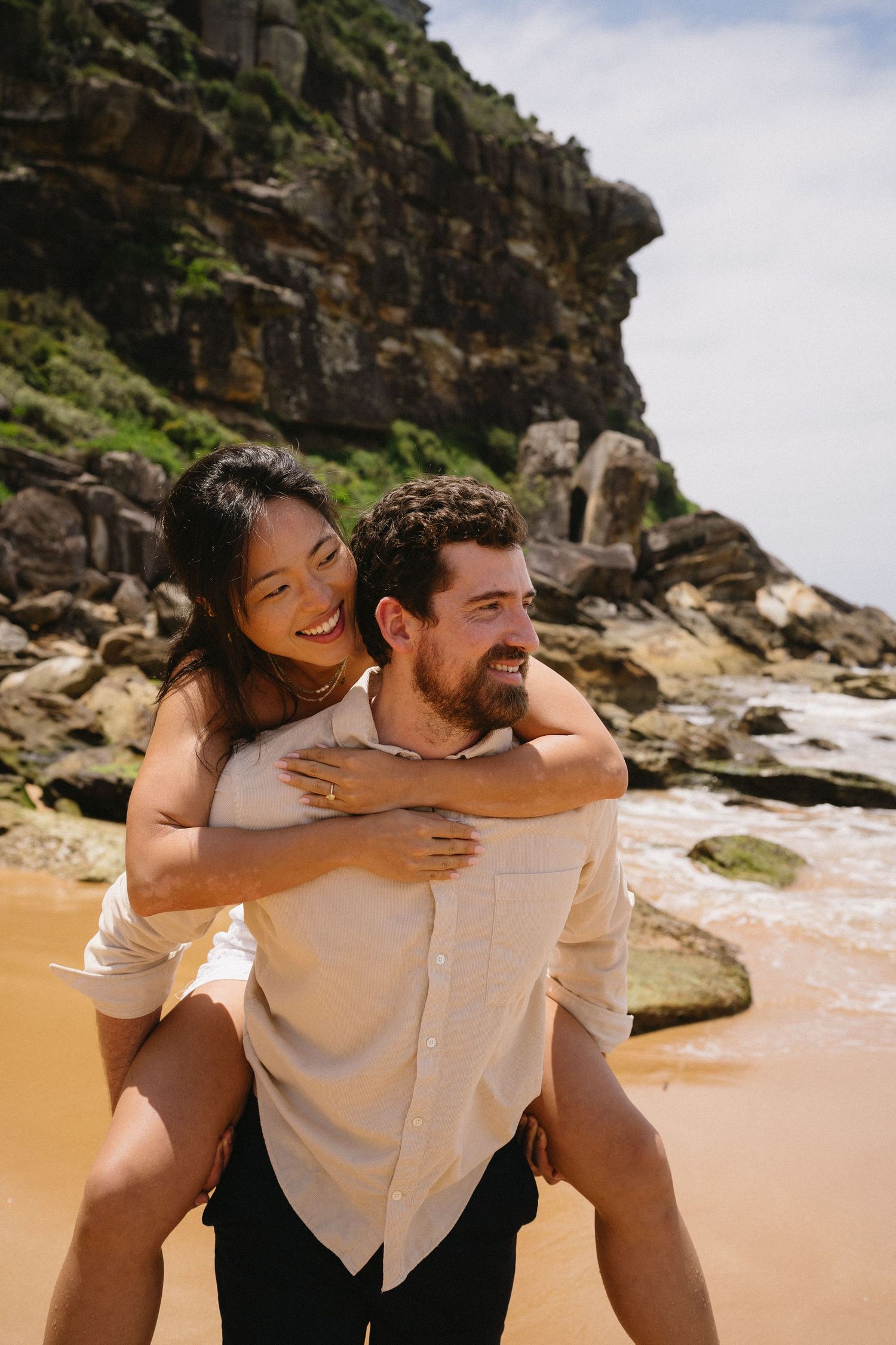 A couple shares a playful piggyback ride moment on a scenic beach with rocky cliffs.