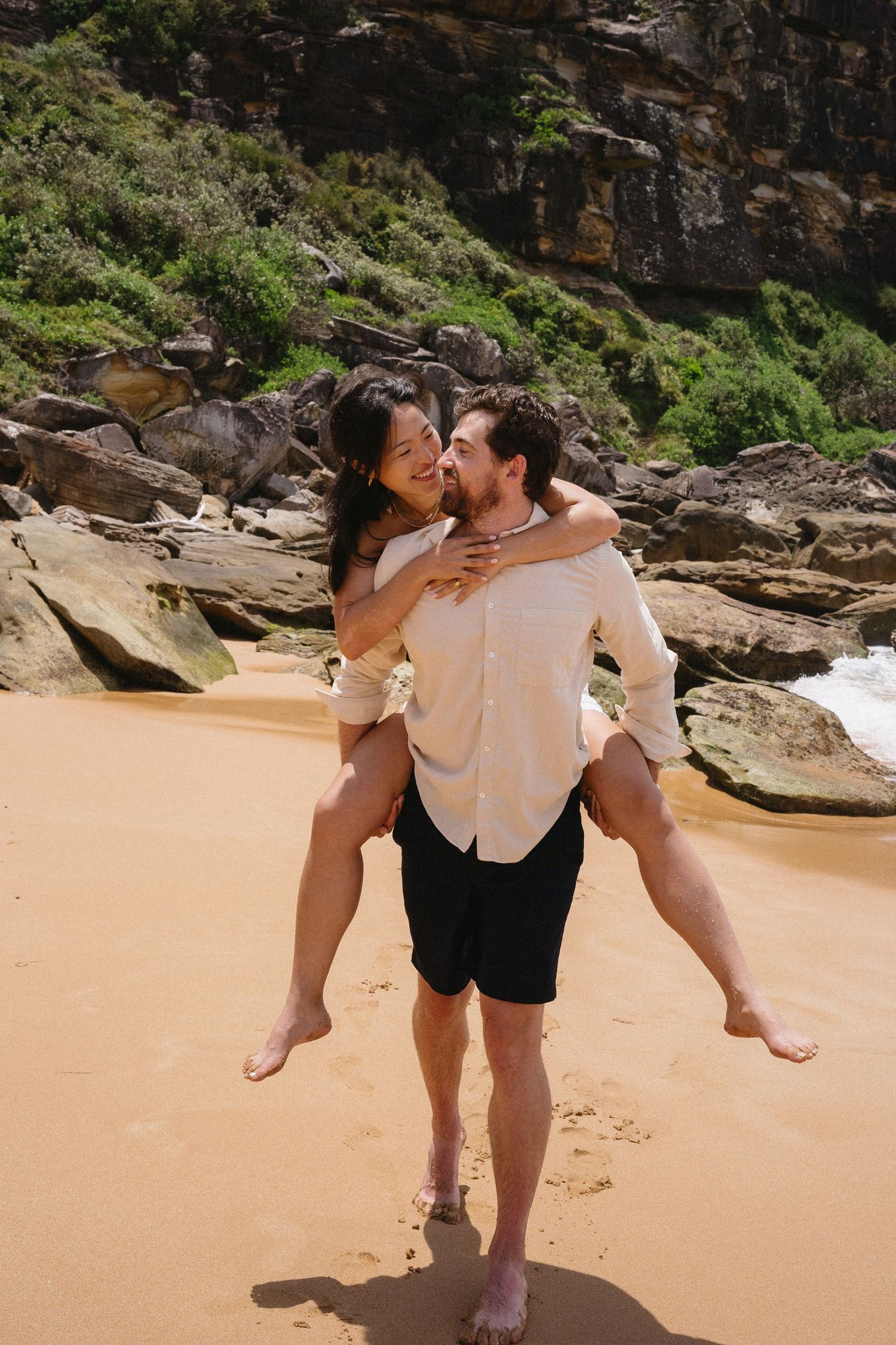 A couple enjoys a piggyback ride moment on a sandy beach with rocky cliffs behind them.