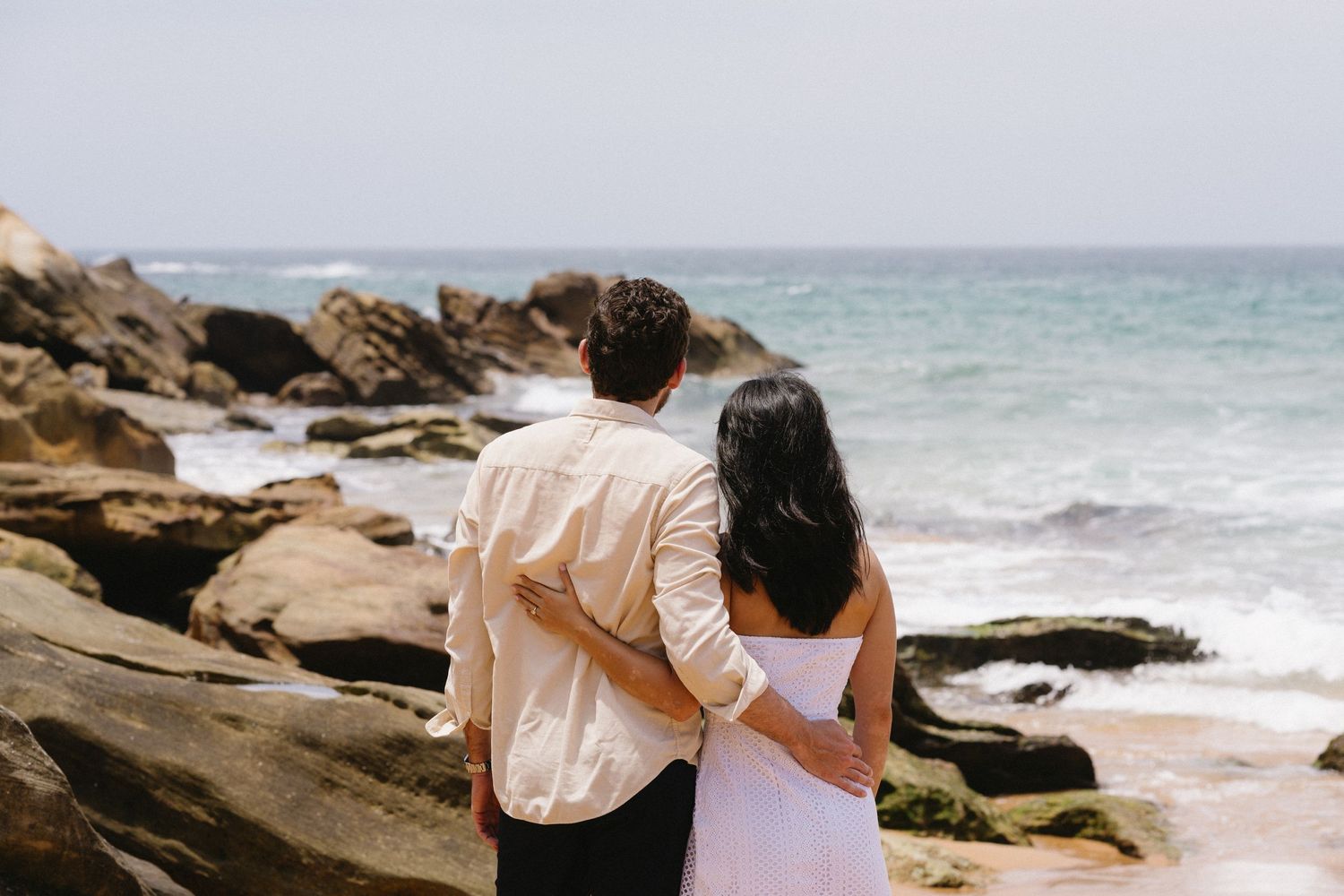 Couple standing arm in arm looking out at ocean waves and rocky shoreline.