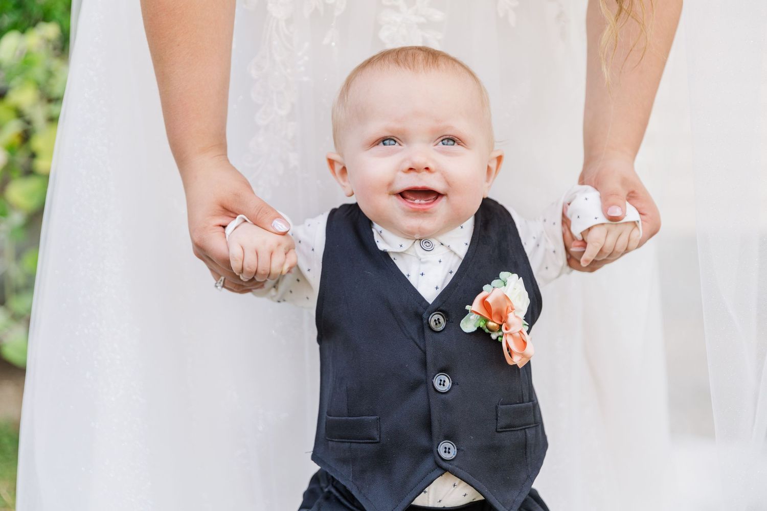A baby in formal vest attire smiles while being held up in a white dress.