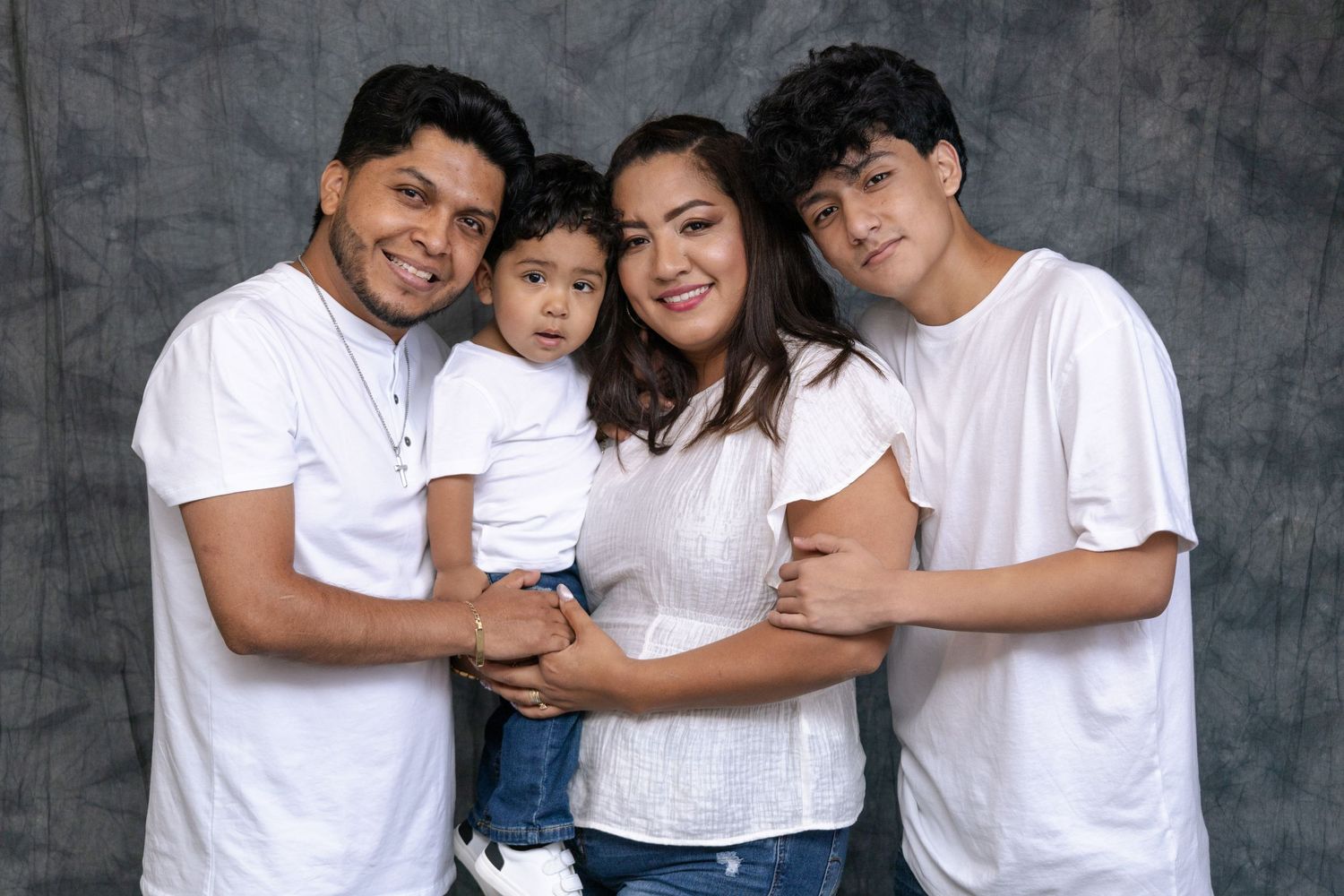 Family portrait session with coordinated white outfits against a textured grey background.