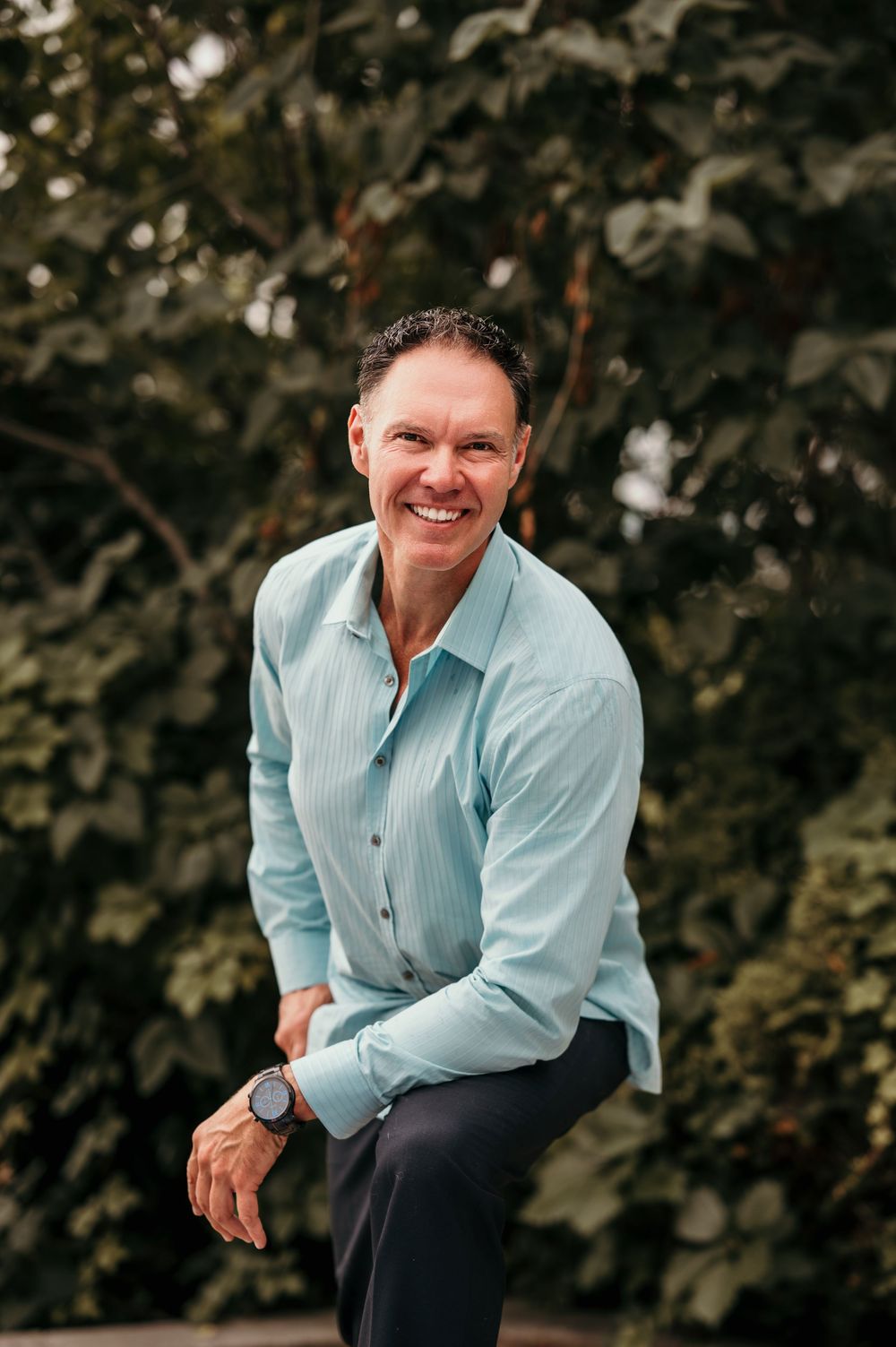 Professional outdoor portrait of someone wearing a light blue button-down shirt against a natural green background.