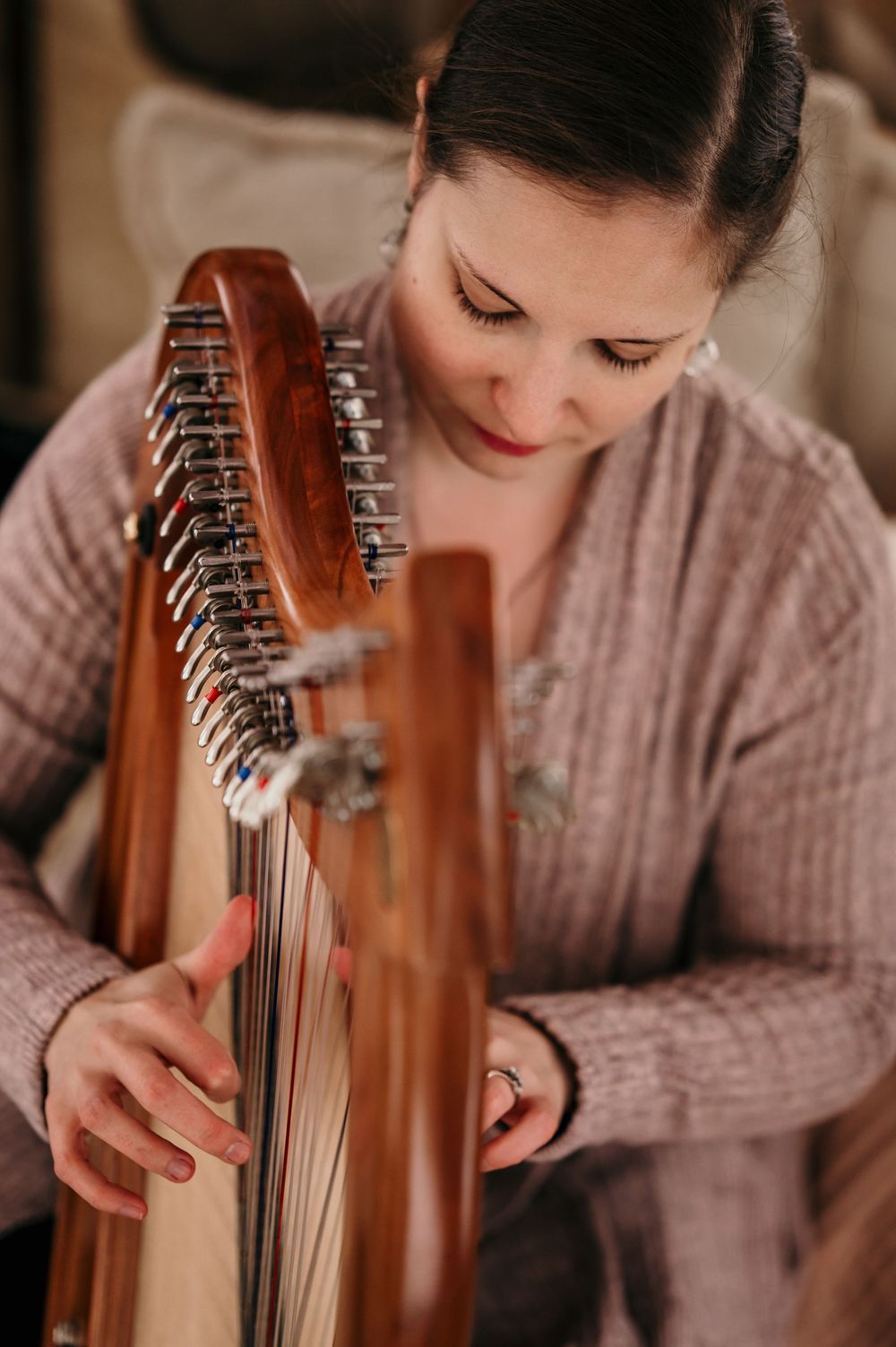 Woman playing a wooden lap harp, focused on strings, wearing a pink sweater in a cozy indoor setting.