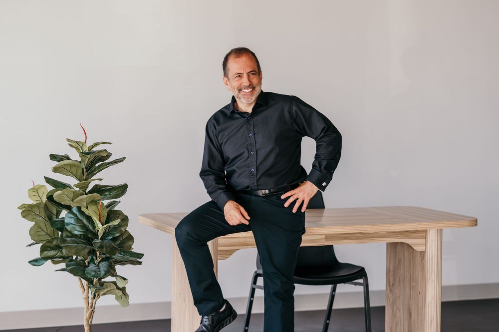Professional man in black shirt smiling, leaning against a wooden table in a modern minimalist office setting.