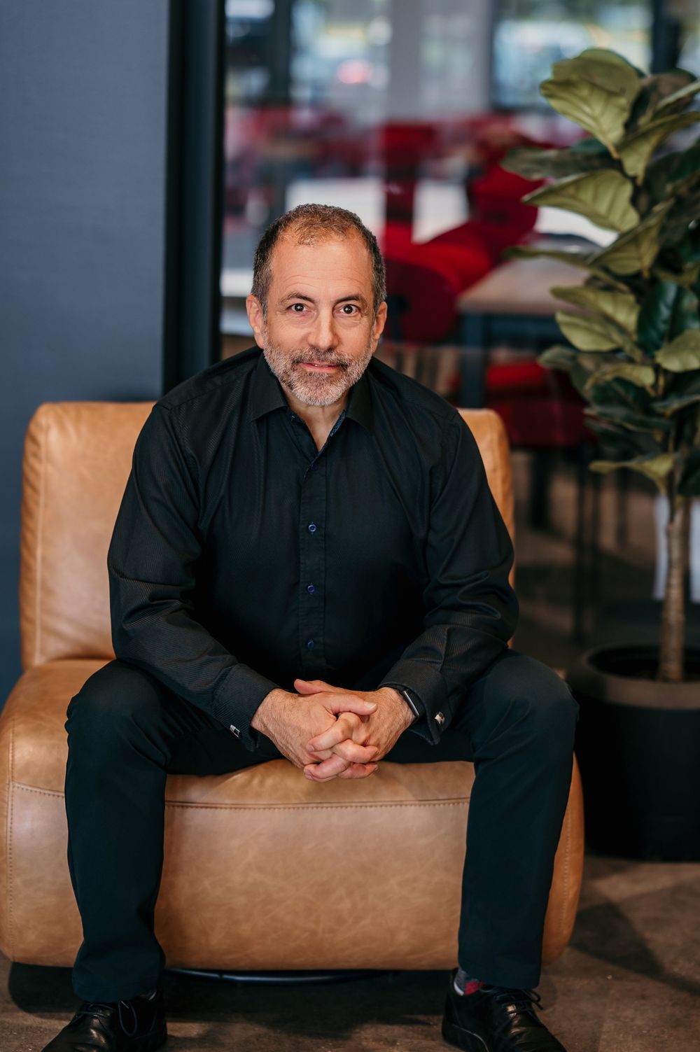 A confident middle-aged man in a black shirt sits in a tan leather chair, smiling with hands clasped.