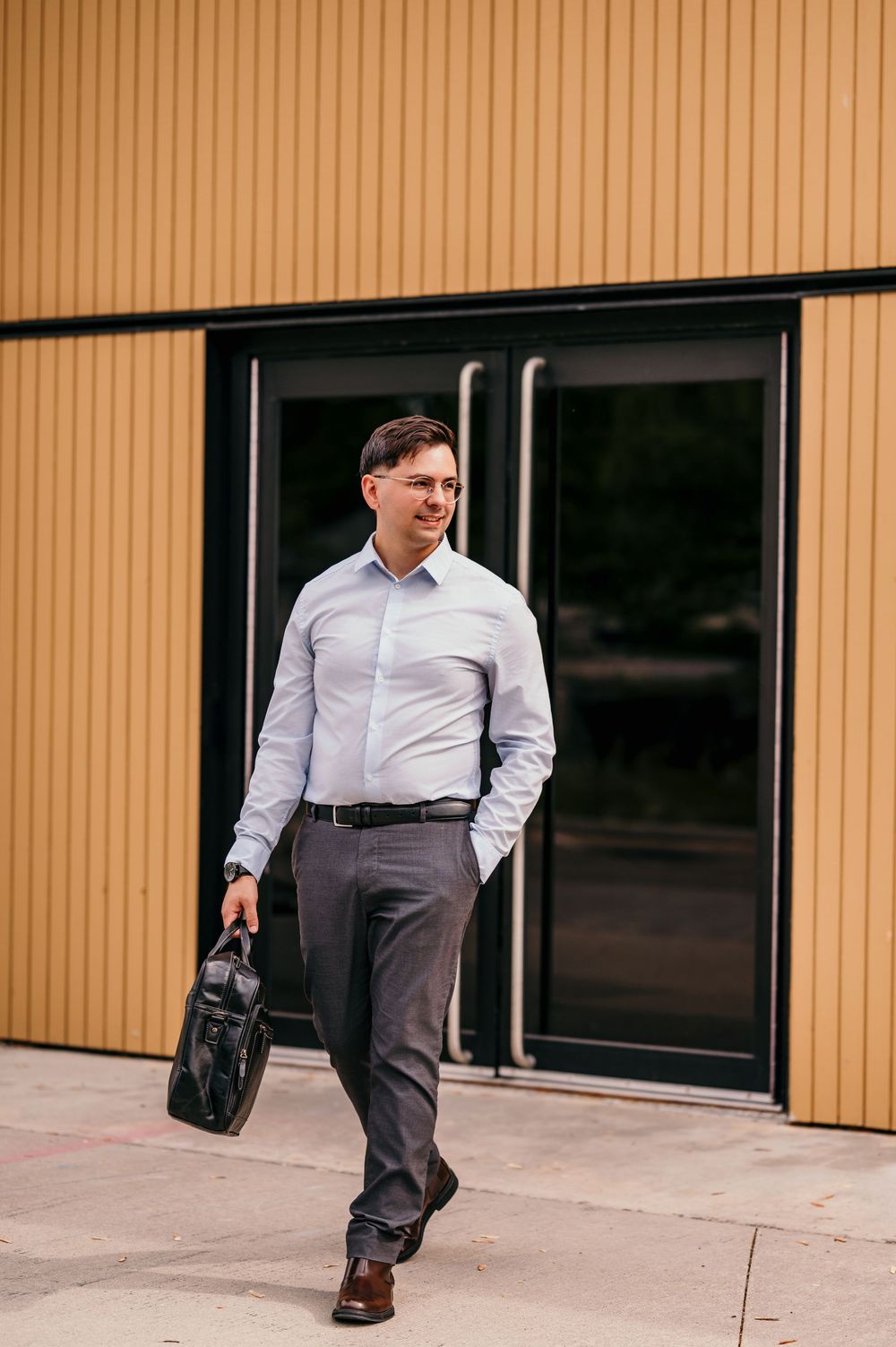 Smiling businessman in white shirt and gray trousers carrying black briefcase walking outside modern yellow building.