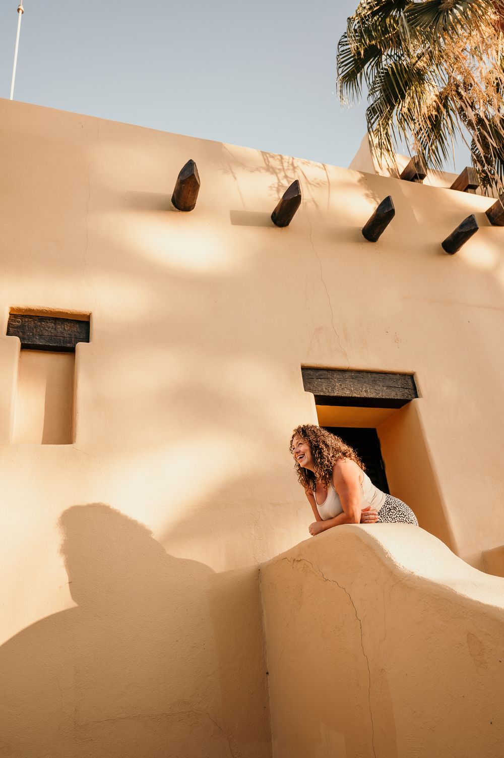 A bride in a white dress leans over an adobe-style wall with vigas and palm trees in the warm desert sunlight.