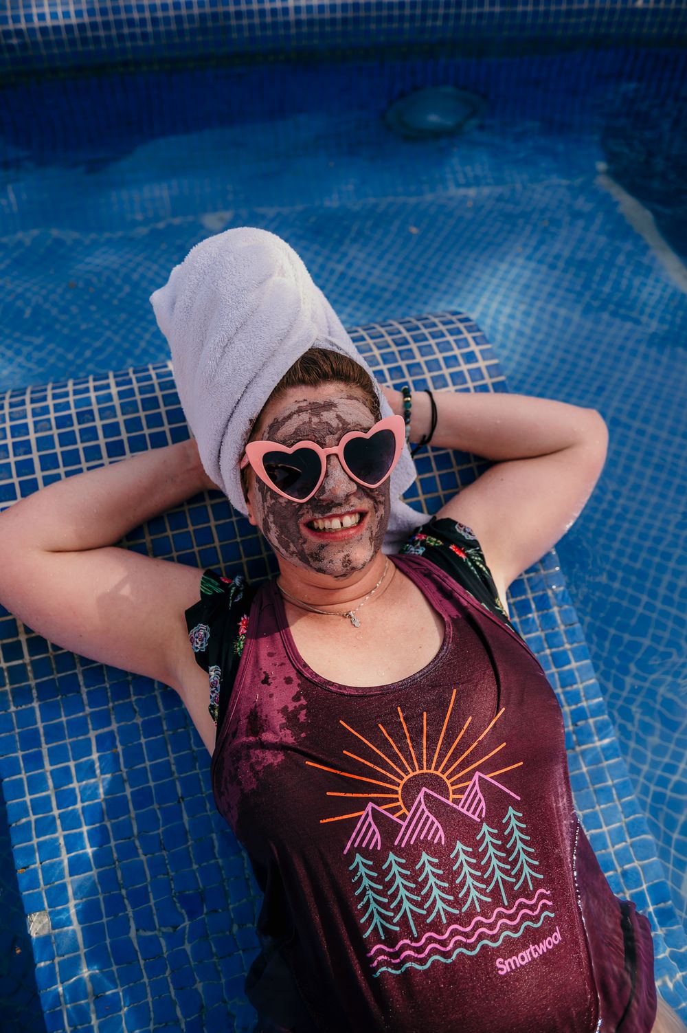 Person in burgundy mountain graphic t-shirt and heart sunglasses relaxing on blue tiled surface with towel wrapped around head.