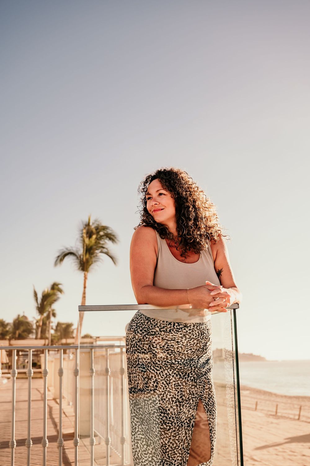 Woman in gray tank top and leopard print skirt poses against railing at sunset with palm trees in background.