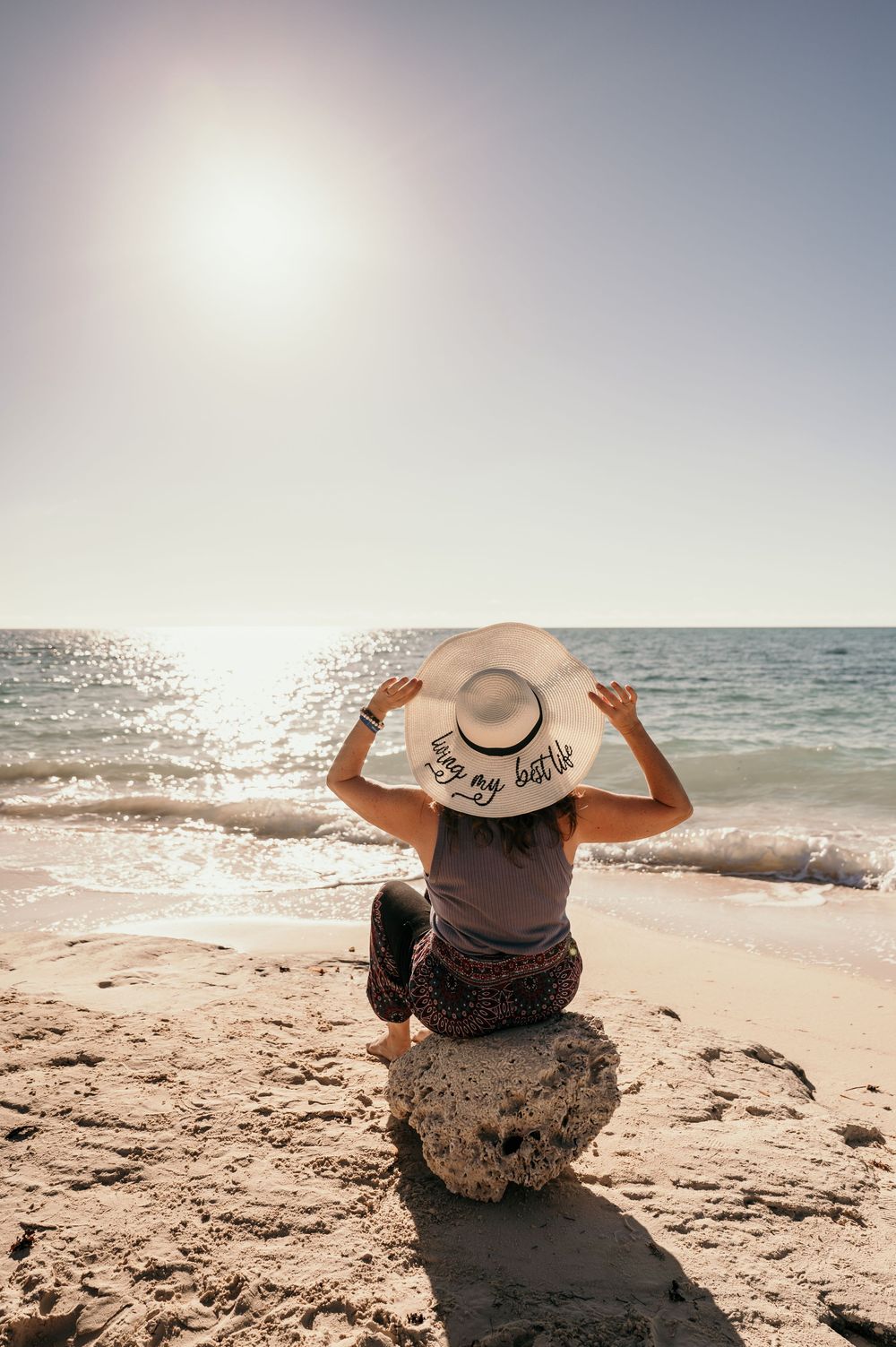 A person wearing a straw sun hat sits on a sandy beach while watching waves roll in at sunset.
