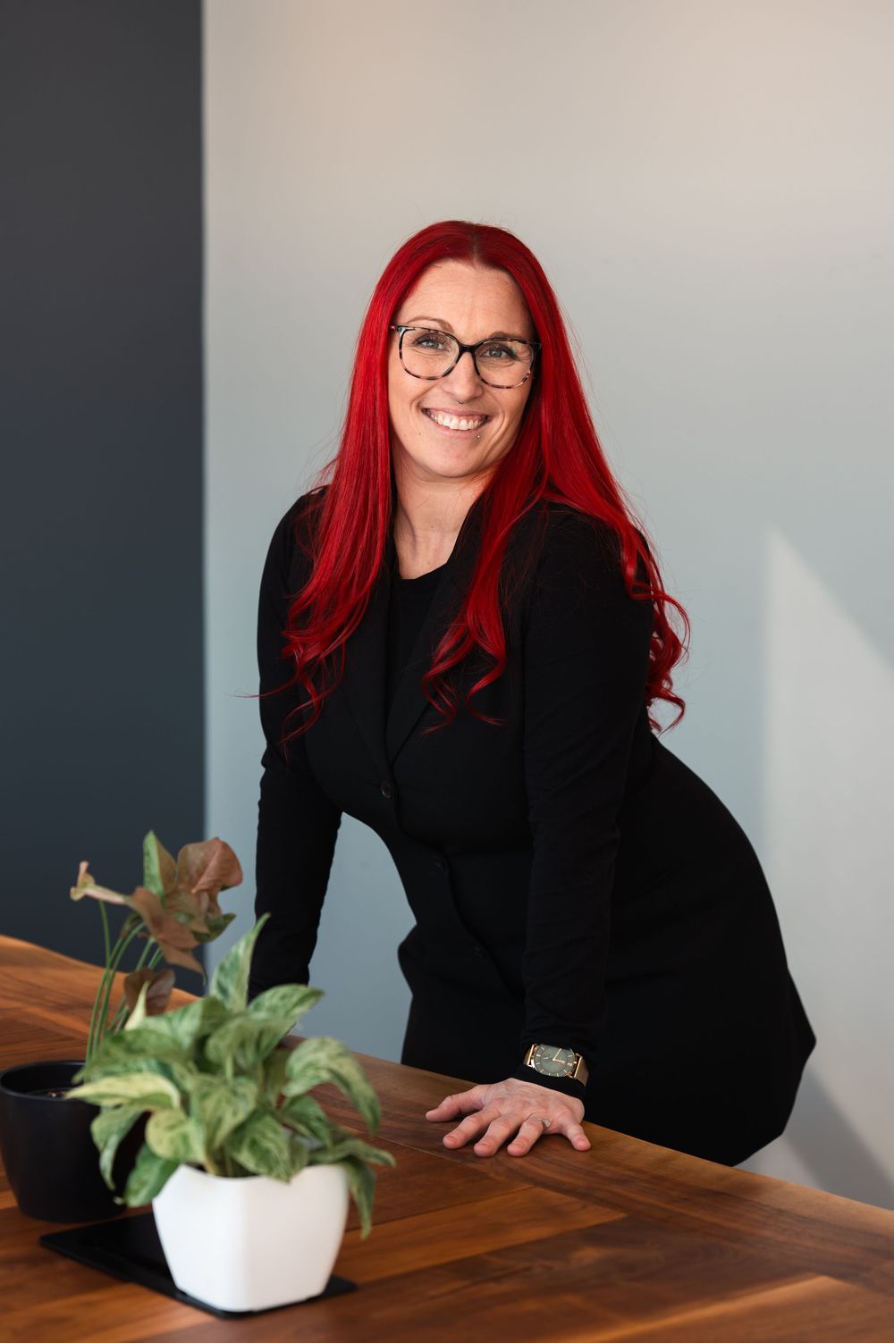 Professional portrait of someone with vibrant red hair wearing a black blazer leaning on a wooden desk with a small plant.