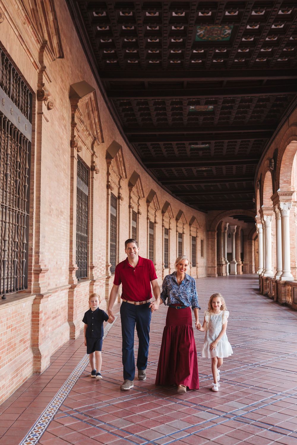 Family of four walking through ornate arched corridor of Plaza de España, Seville, Spain.
