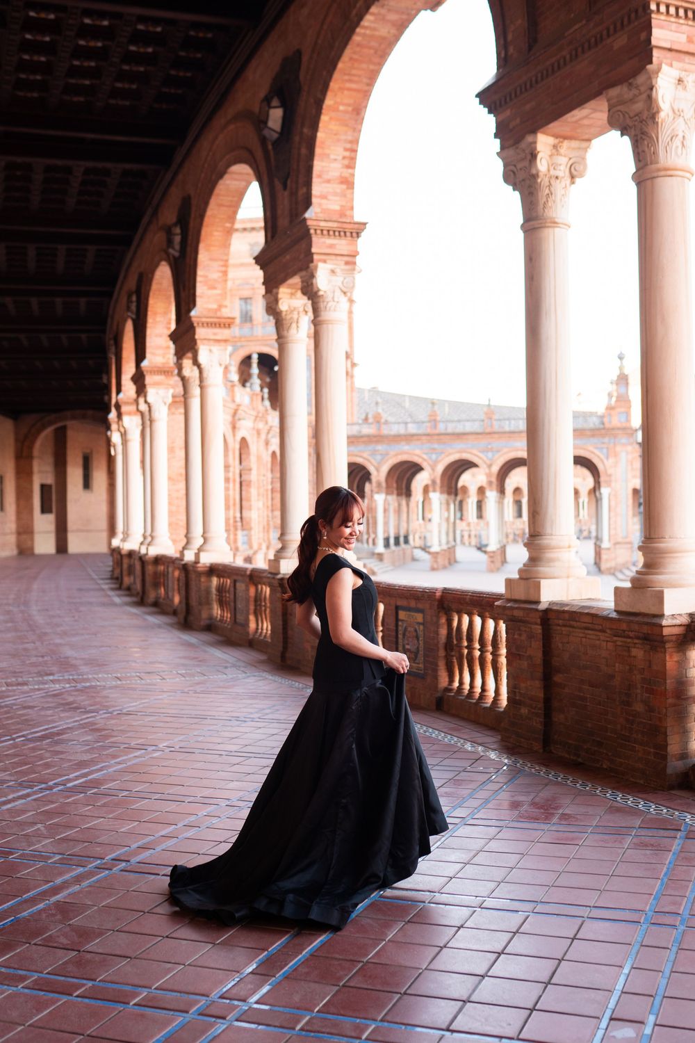 Woman in elegant black gown posing under arched colonnade at Plaza de España, Seville, Spain.