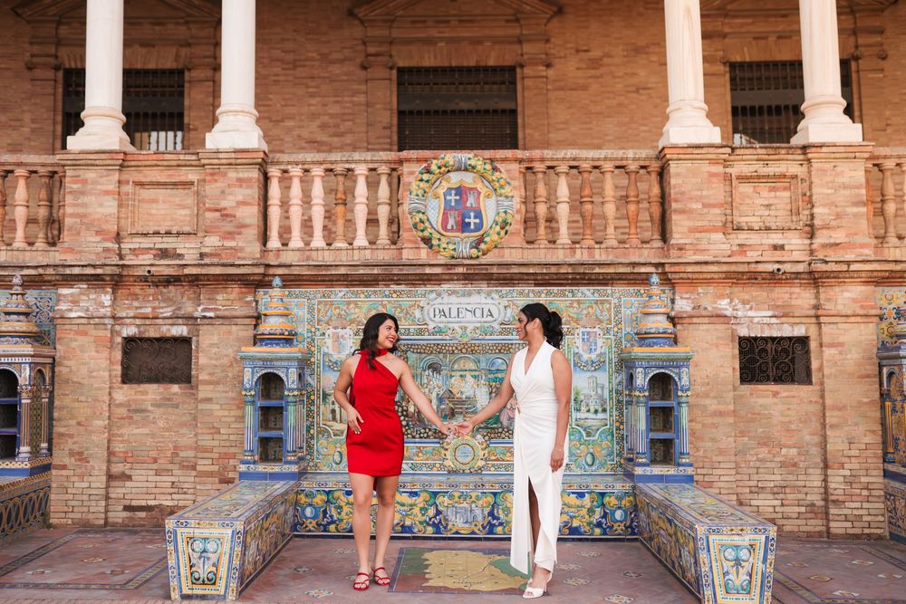 Two women in red and white dresses hold hands at Plaza de España, Seville, with colorful tiled alcoves behind them.