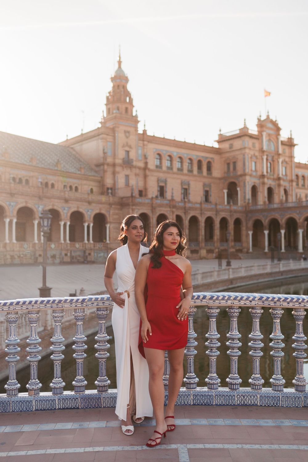 Two women in elegant red and white dresses pose on a bridge at Plaza de España, Seville, Spain.
