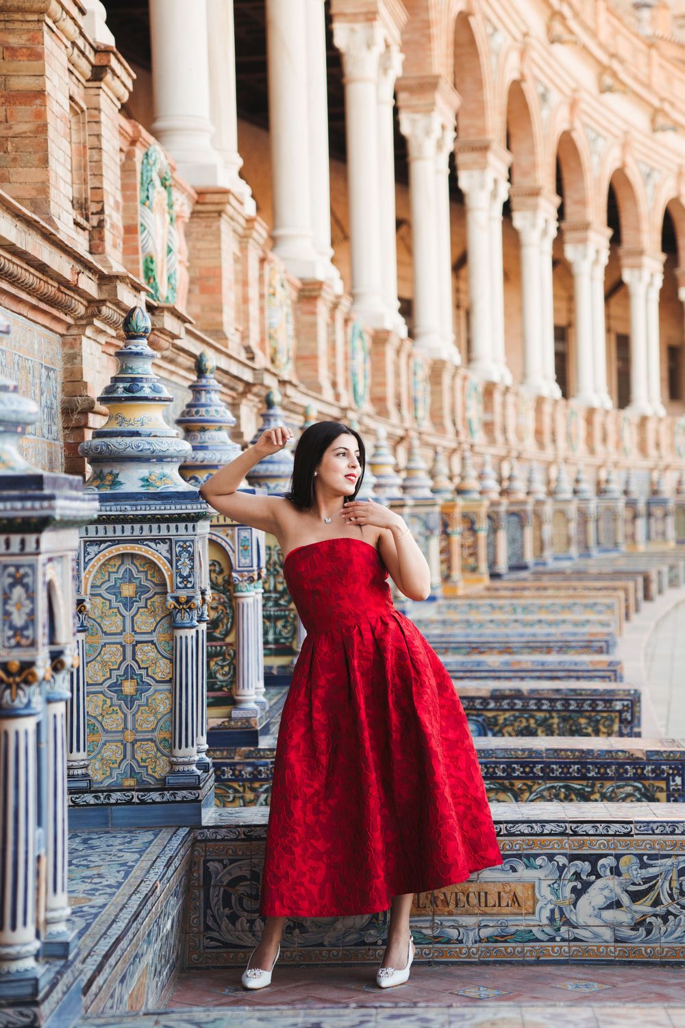 Red dress photoshoot along the ornate tiled colonnade of Plaza de España in Seville.