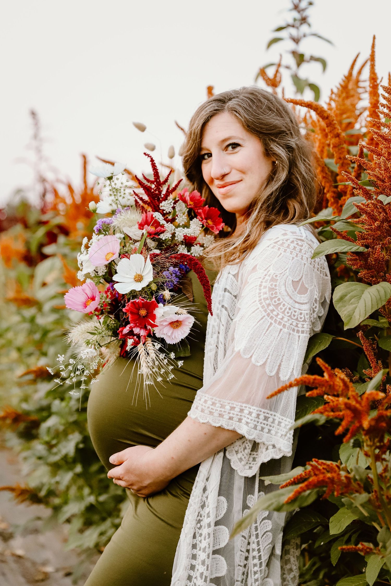 Expectant mother in lace dress holds colorful bouquet against autumn foliage.
