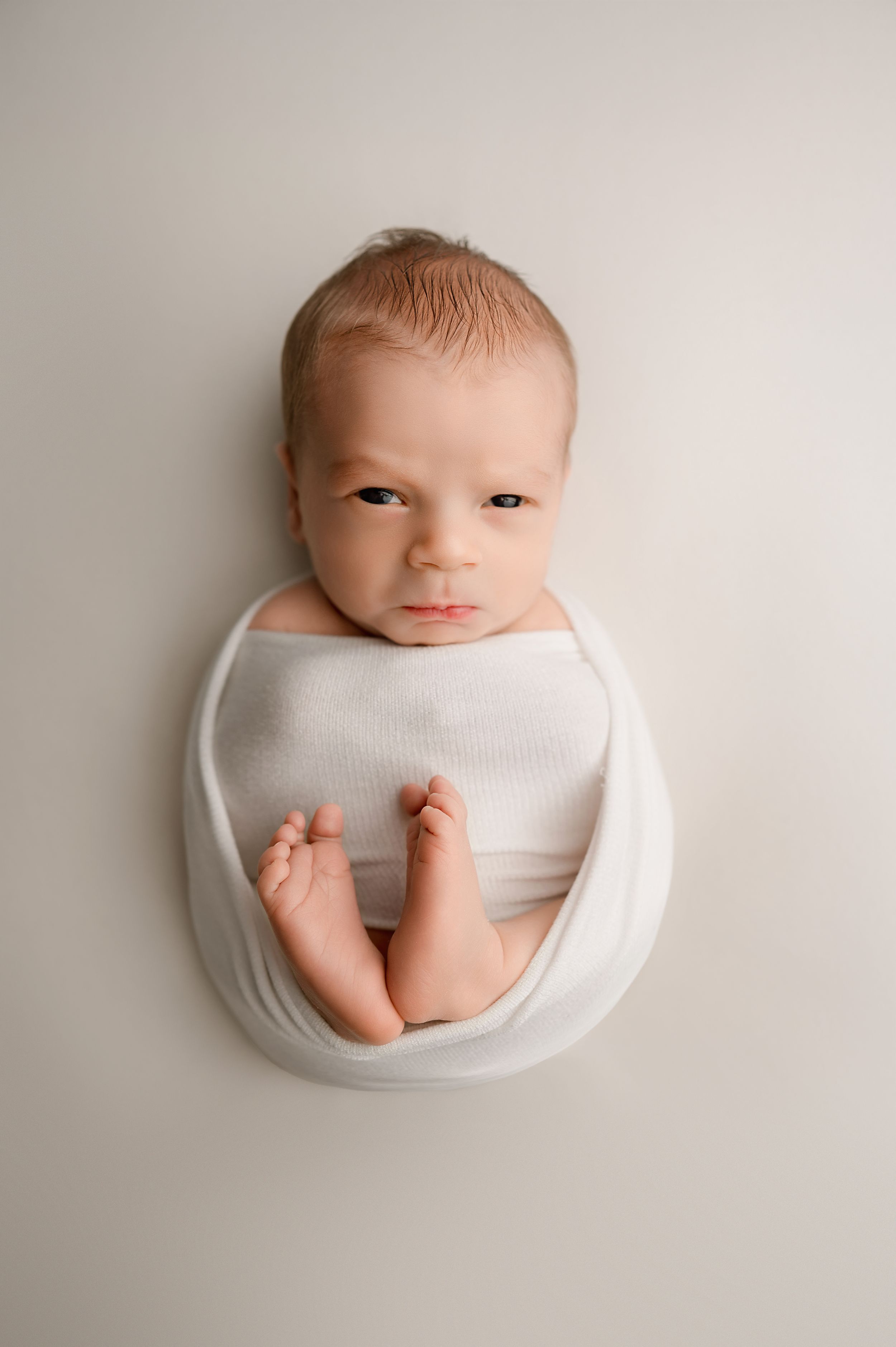 Delicate newborn photography series showing baby wrapped in white fabric against neutral background.