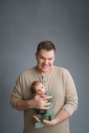 A person in a beige sweater holds and plays with a tiny baby wearing mint green sleepwear against a grey backdrop.