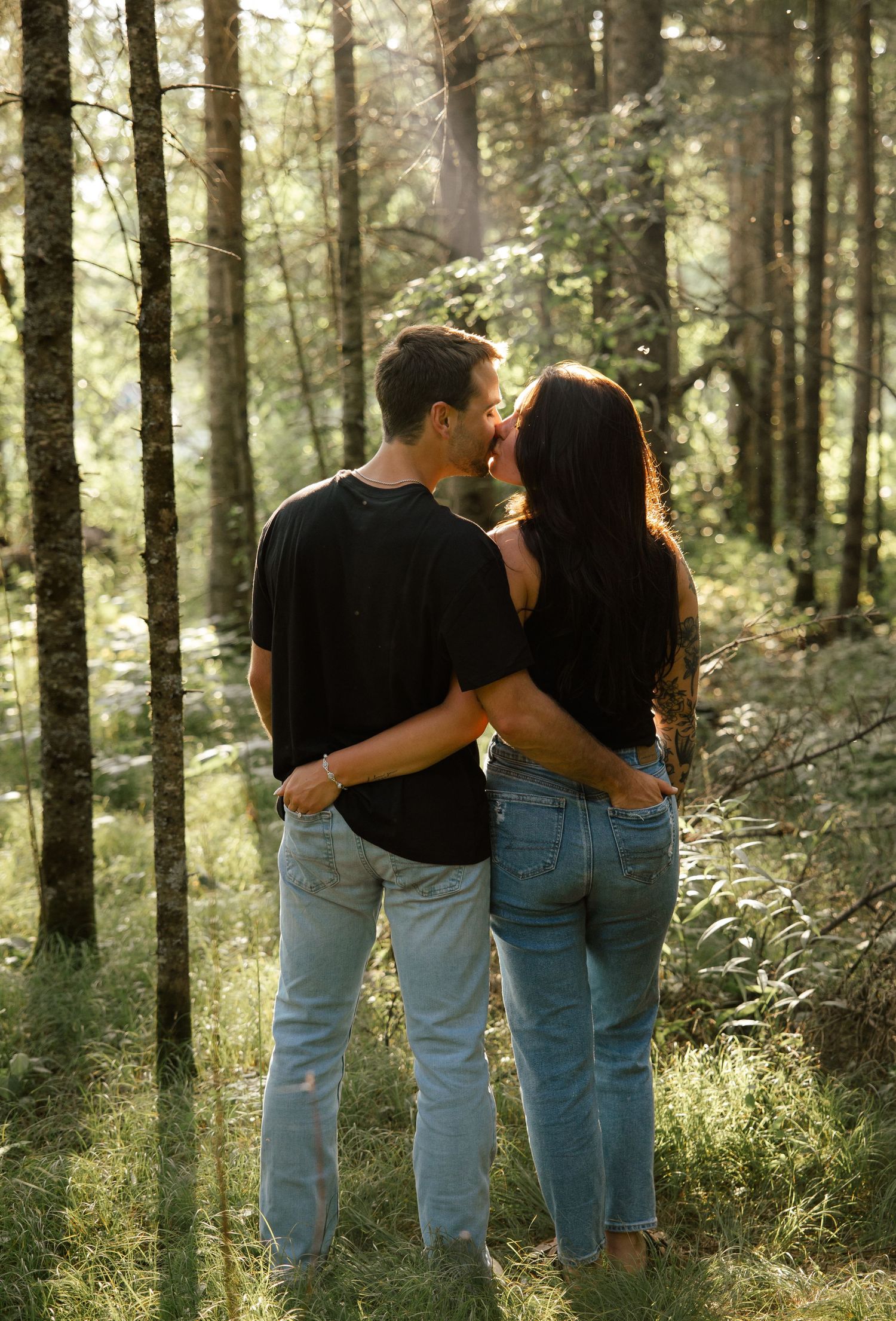 people wearing jeans and black shirts walk together through a sunlit forest path.