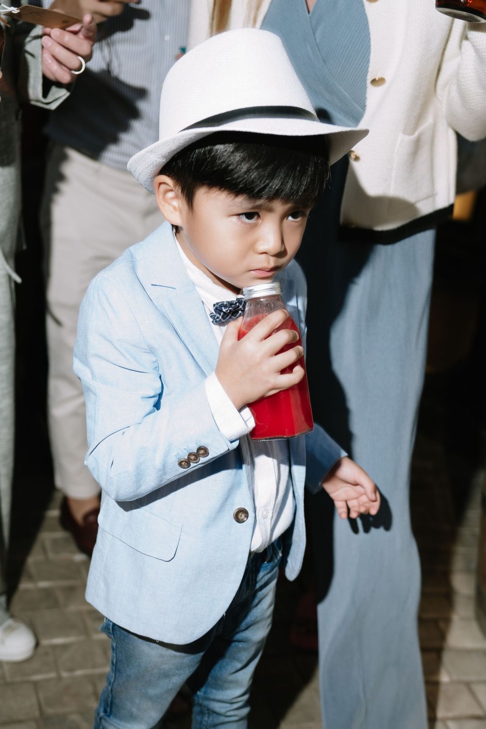 Stylish young boy in white hat and blue blazer drinking red beverage at evening event.