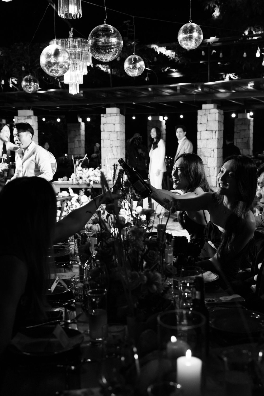 Black and white photo of guests toasting at candlelit wedding reception with disco ball decor.