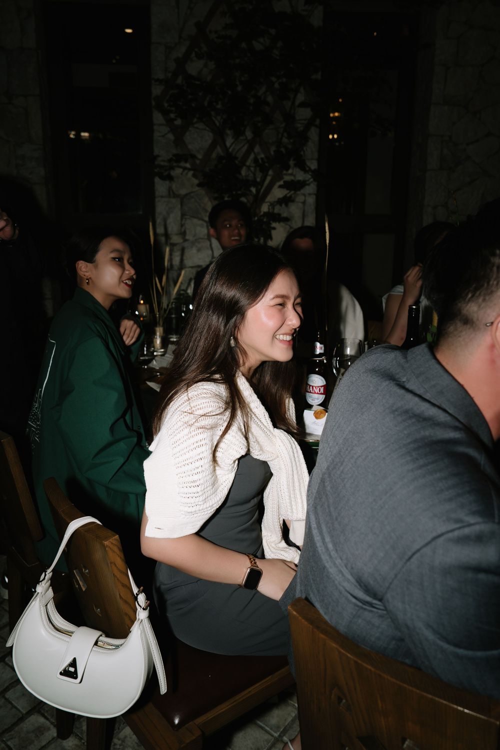 Smiling young woman in white cardigan seated at evening dinner party with designer handbag.