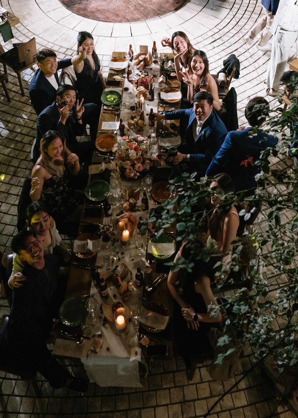 Overhead view of wedding guests smiling and waving at a candlelit long dinner table.