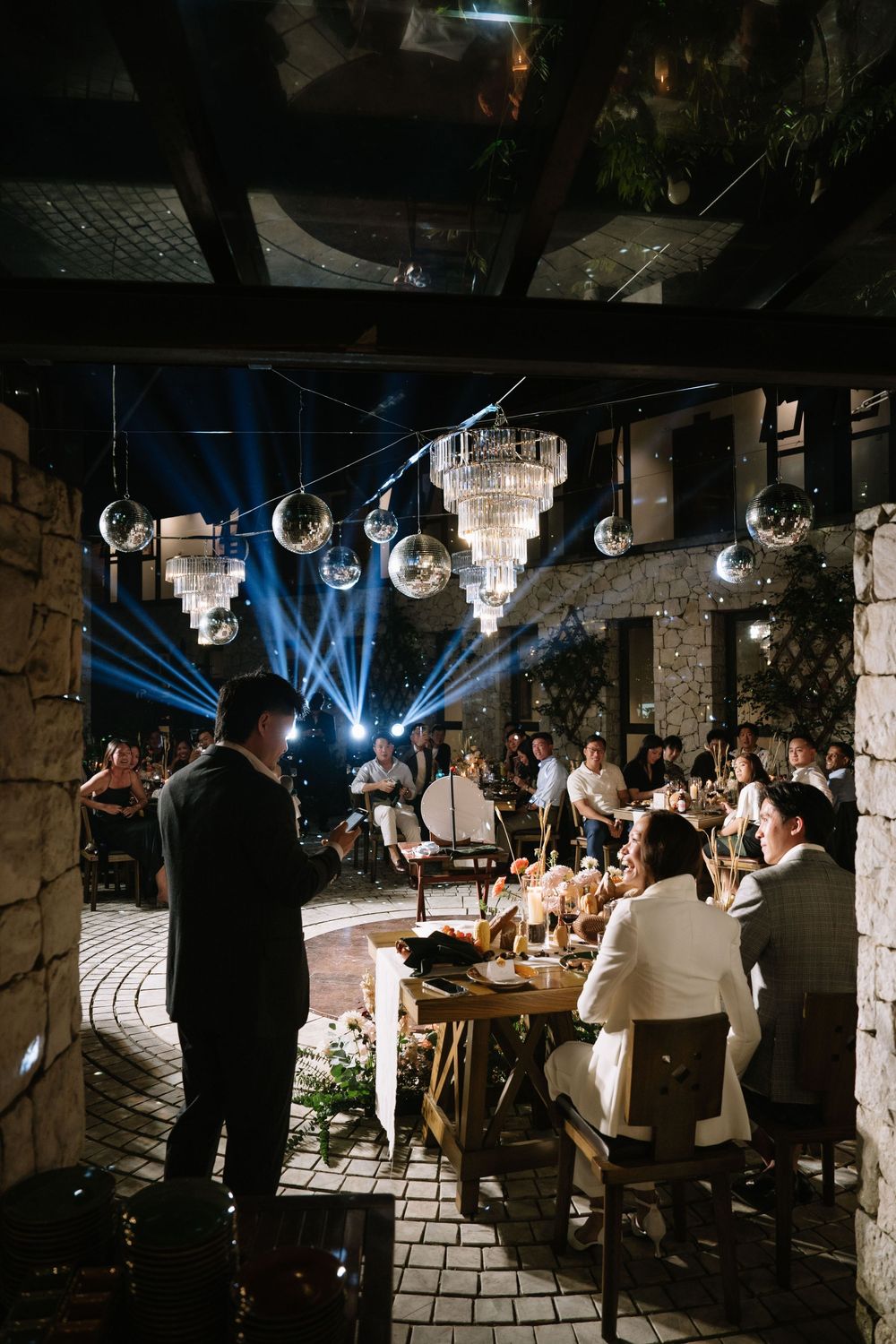 Guests dining at a stylish outdoor wedding reception with crystal chandeliers and disco balls overhead.
