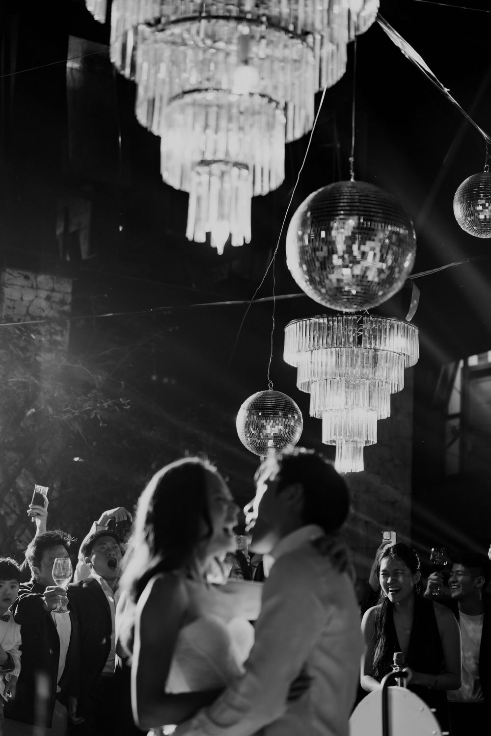 Black and white photo of newlyweds kissing under disco balls and chandeliers at their reception.