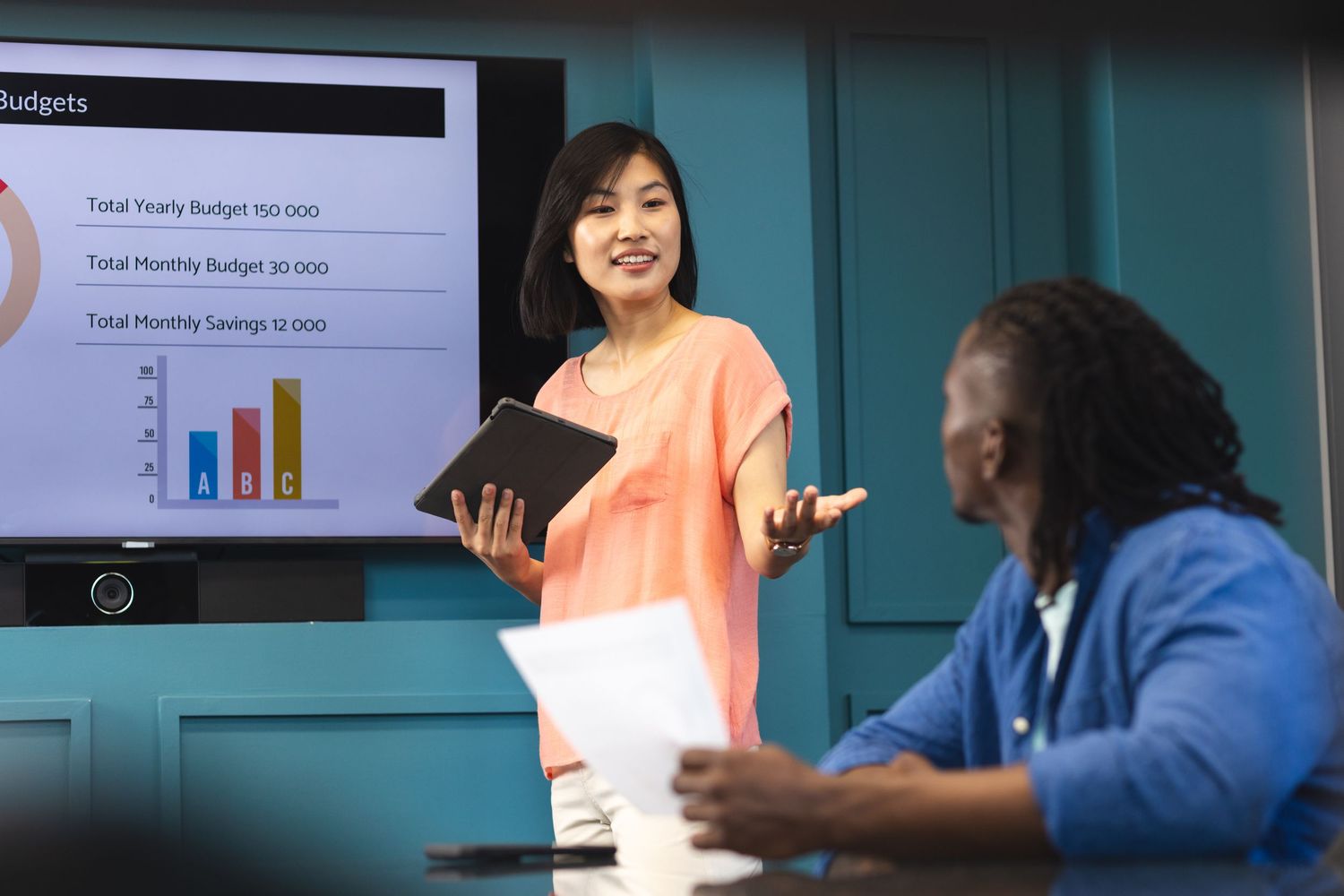A presenter explains financial data and charts during a business meeting while gesturing at a projection screen.