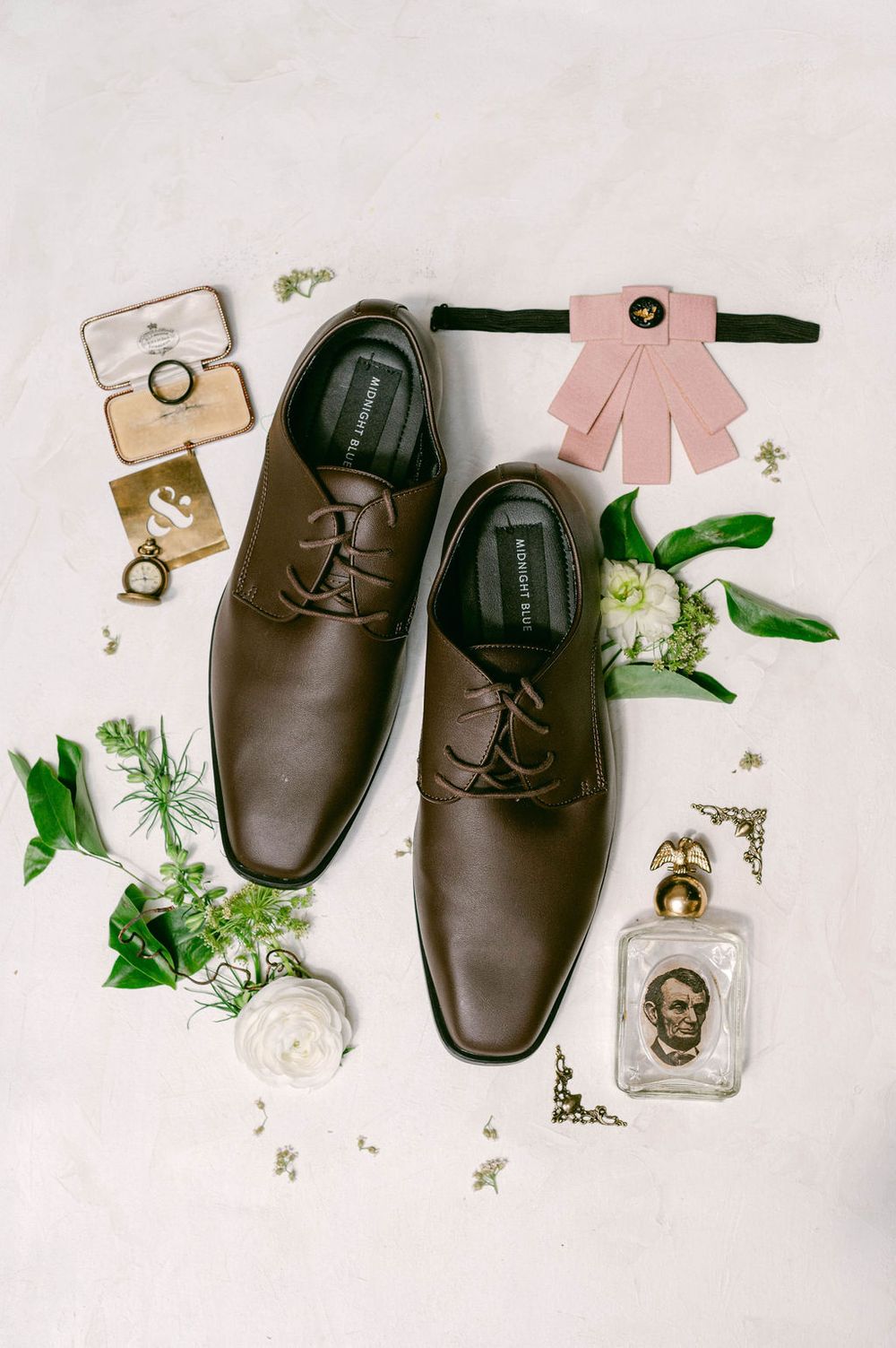 Brown leather dress shoes arranged with delicate white flowers and greenery on white background for wedding photography.