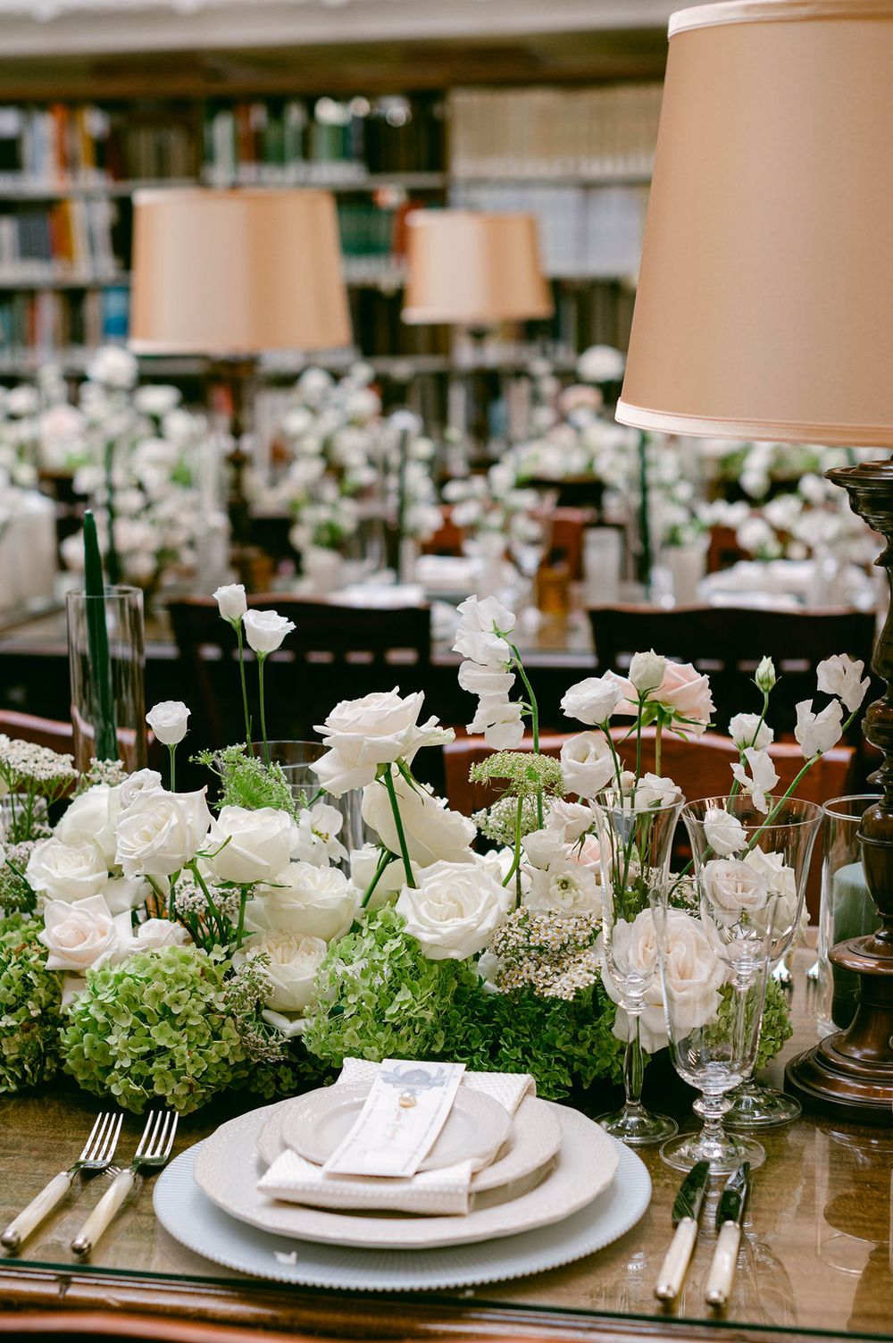 Elegant wedding table setting with white roses, greenery, and fine china on a wooden table with ambient lighting.