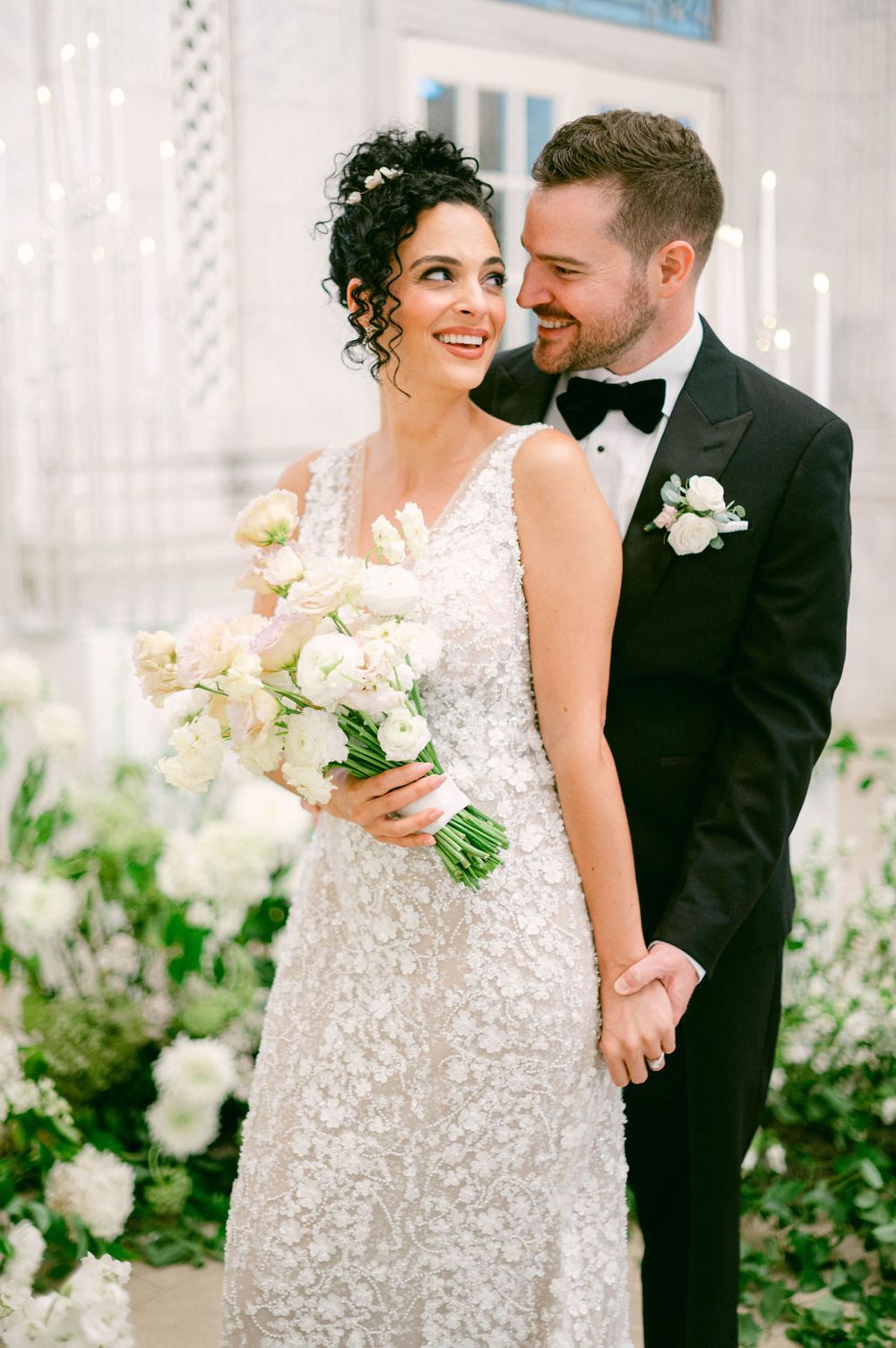 Romantic wedding portrait shows a couple surrounded by lush white floral arrangements in an elegant outdoor setting.