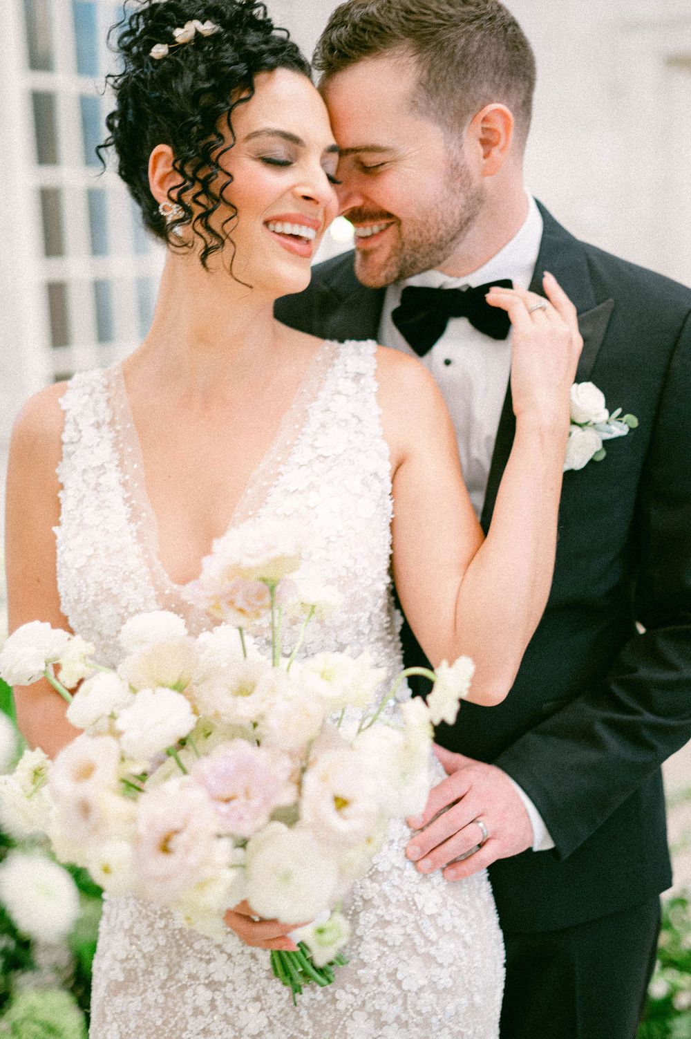 Romantic wedding couple embrace while holding white floral bouquet in an elegant outdoor setting.