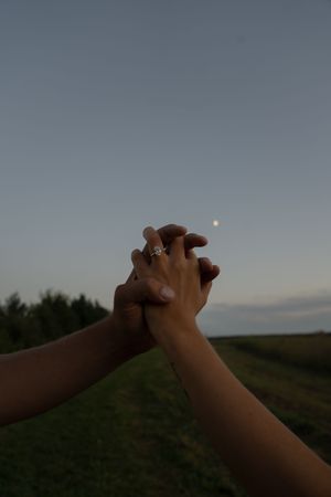 Two hands clasp together against a twilight sky with the moon visible in the background.
