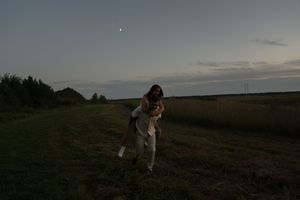 A couple embraces in a grassy field at dusk with trees in the background.