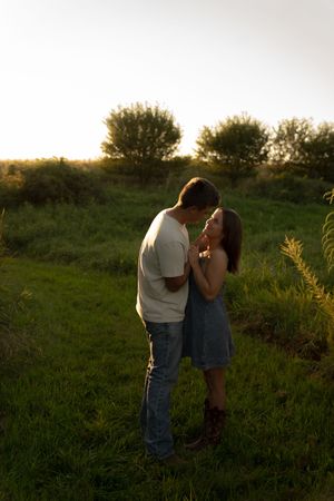 A romantic couple stands close together in a field during golden hour.