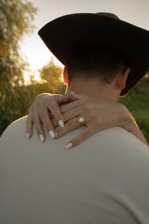 Close-up of hands displaying an engagement ring against a white shirt and cowboy hat at sunset.