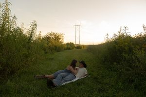 Couple lying on a blanket in a rural field at sunset with power lines in the distance.
