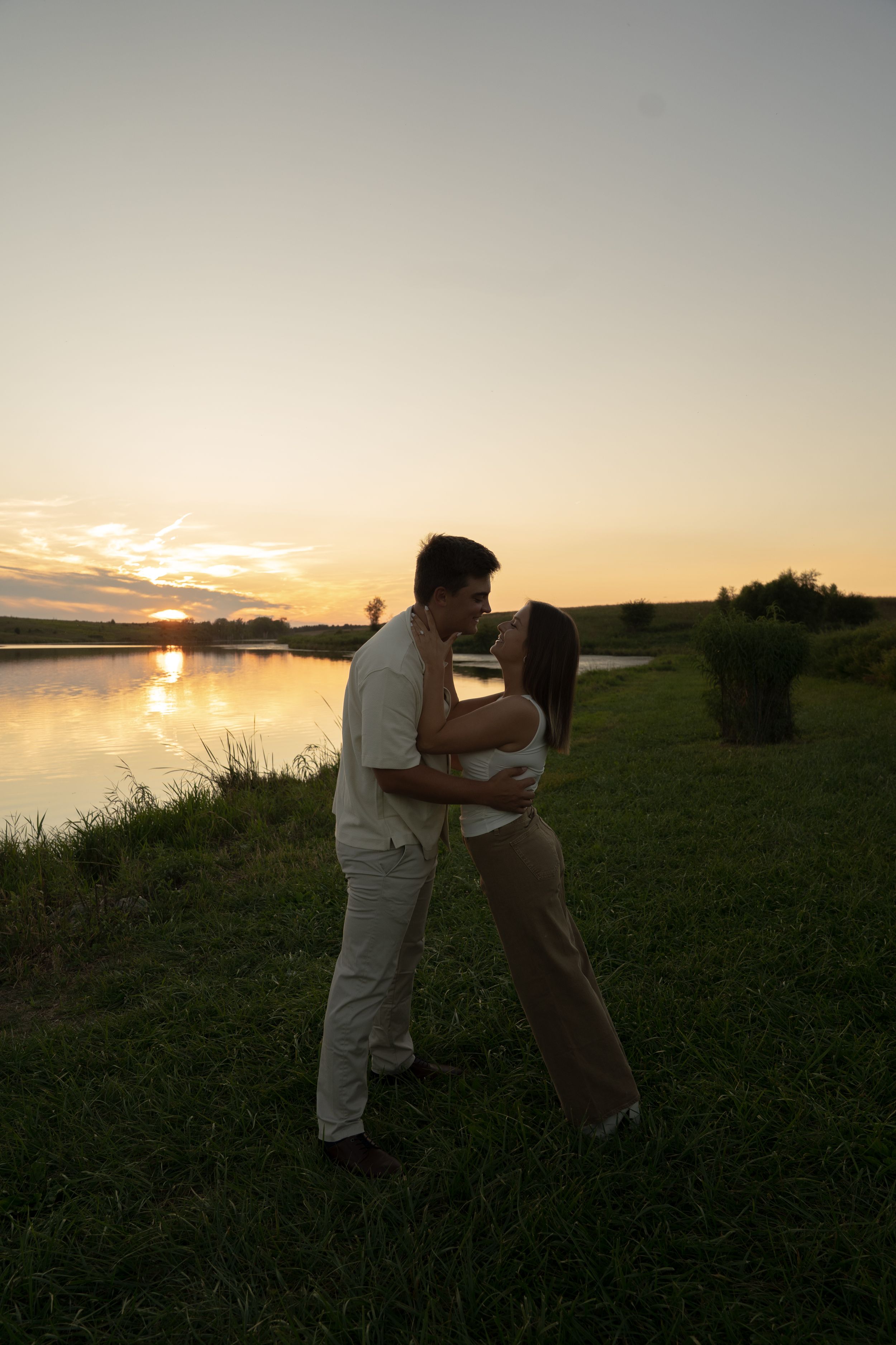 Two people embrace on a grassy hillside overlooking a serene lake at sunset.