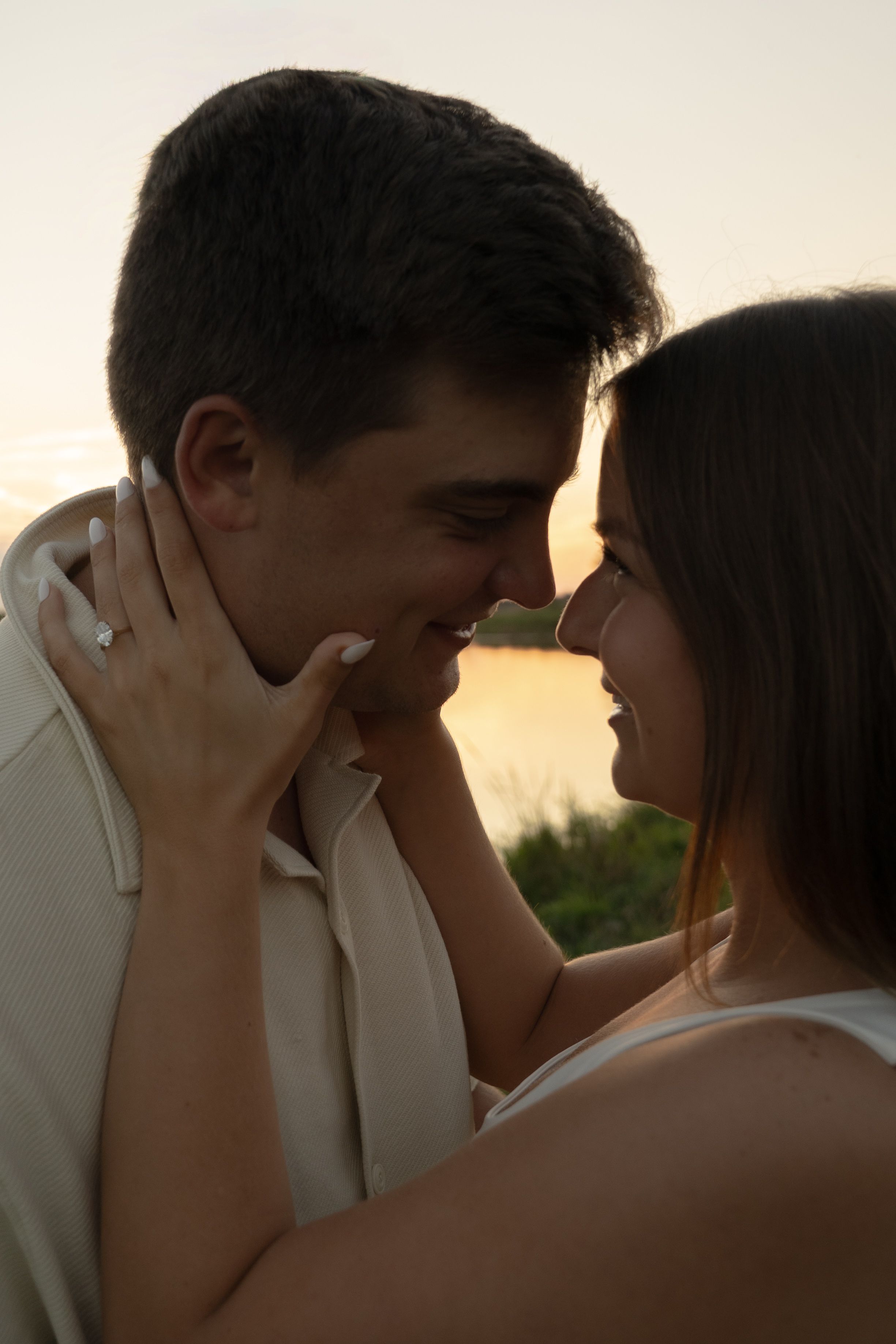 A romantic couple embraces against a glowing sunset backdrop near a lake.