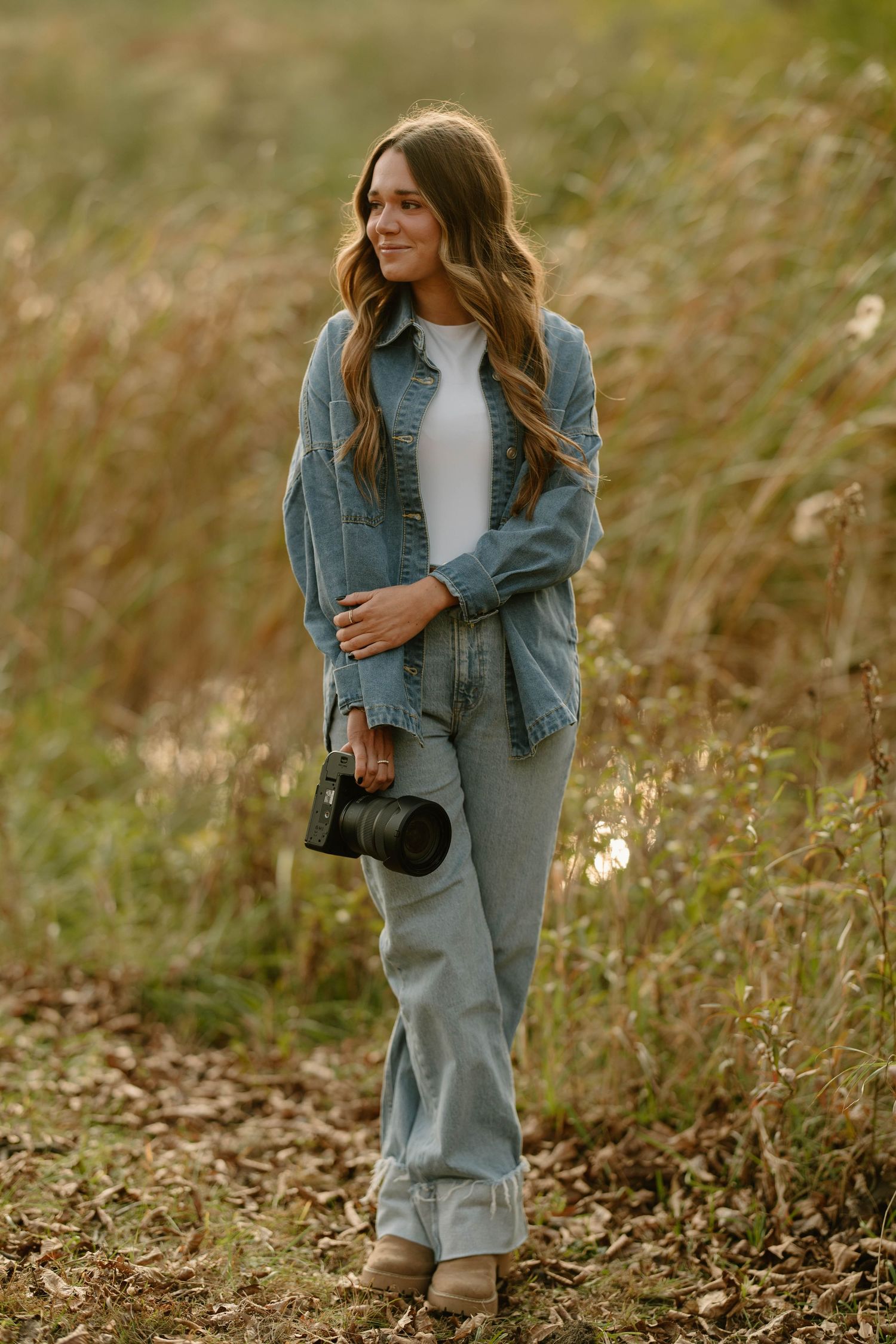 Person in denim jacket walking through tall grass during golden hour.