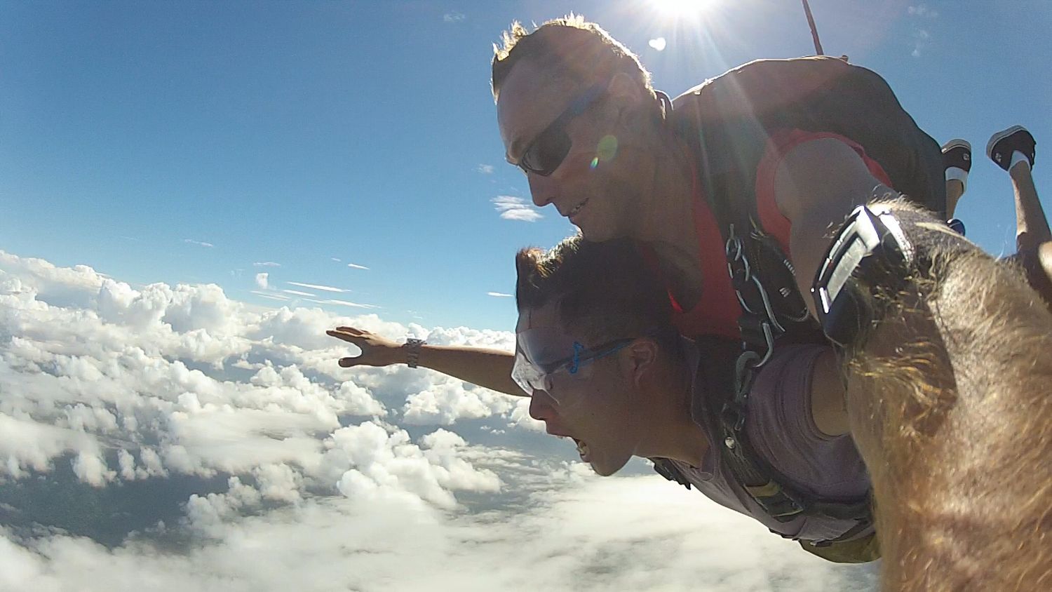 Skydiving tandem pair soar through bright blue skies above fluffy white clouds during thrilling freefall descent.
