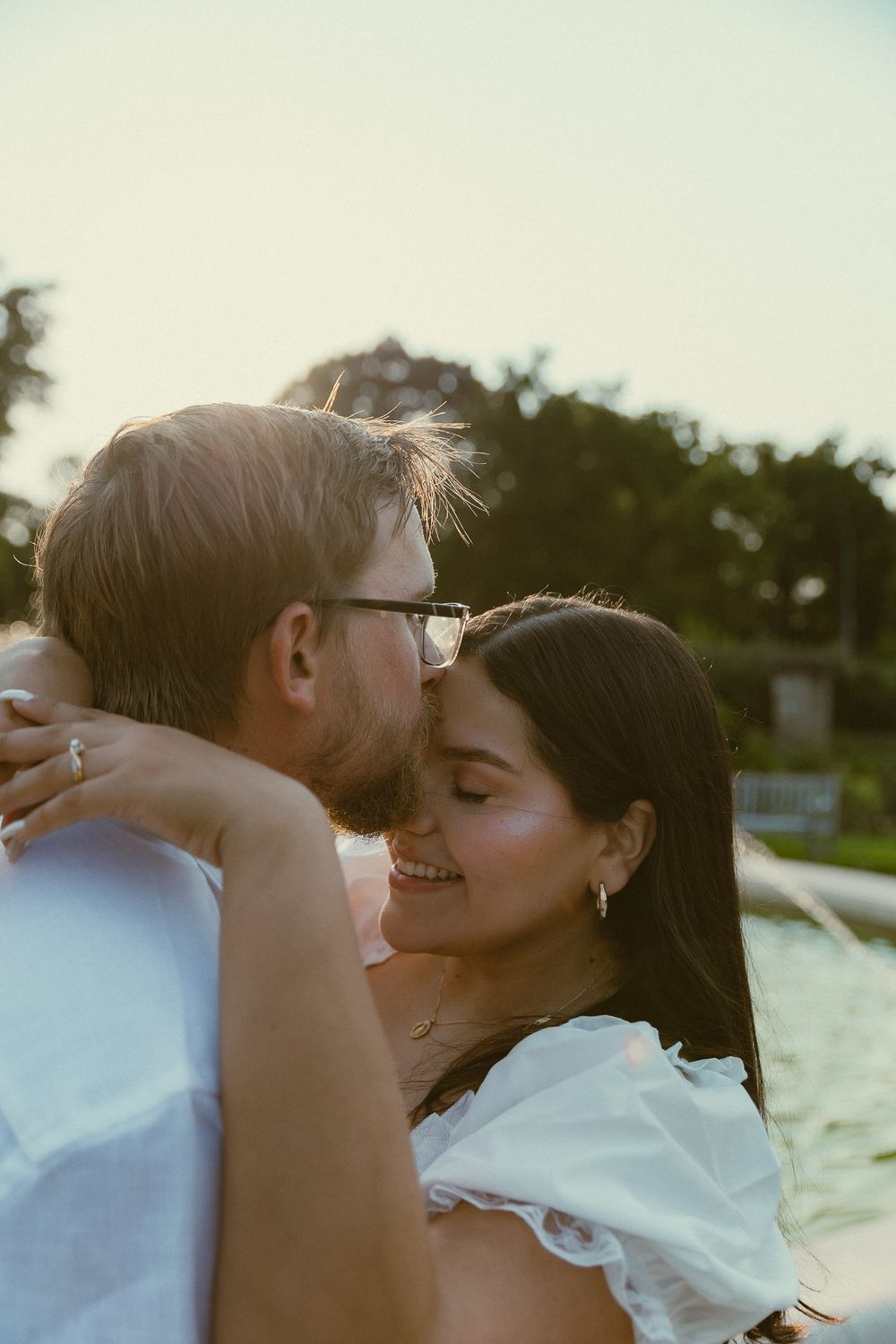 A couple shares a tender romantic moment outdoors at golden hour, the woman smiling as the man leans in to kiss her cheek.