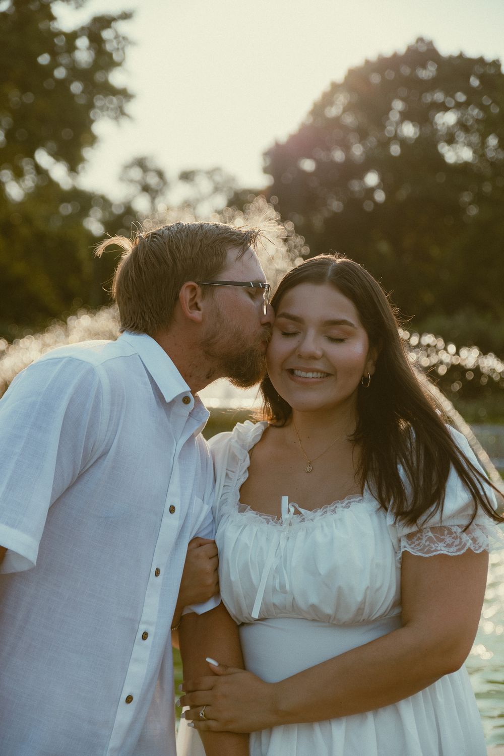 A couple shares a tender moment outdoors at golden hour, man kissing womans cheek as she smiles in a white dress.