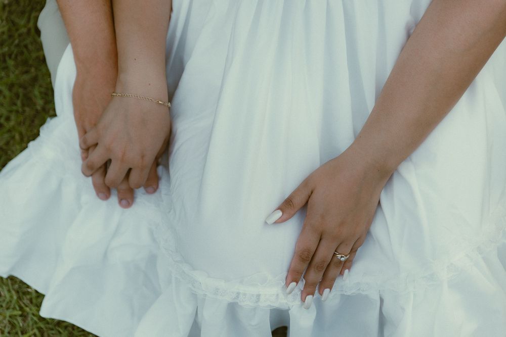 Two women in white dresses holding hands with gold jewelry and white manicured nails outdoors.