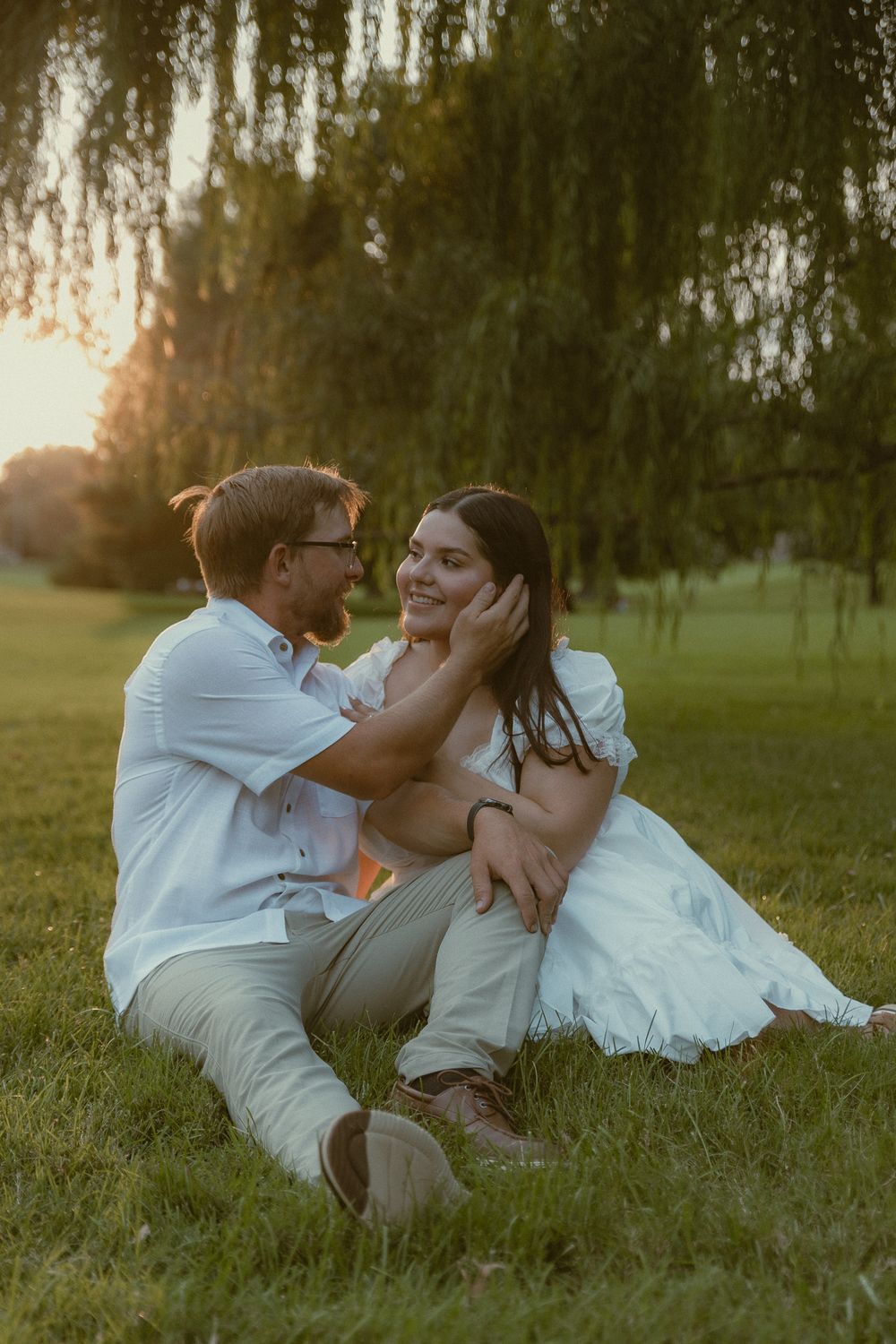 Couple sitting on grass under willow tree at golden hour, man tenderly cupping woman's face as they gaze at each other.