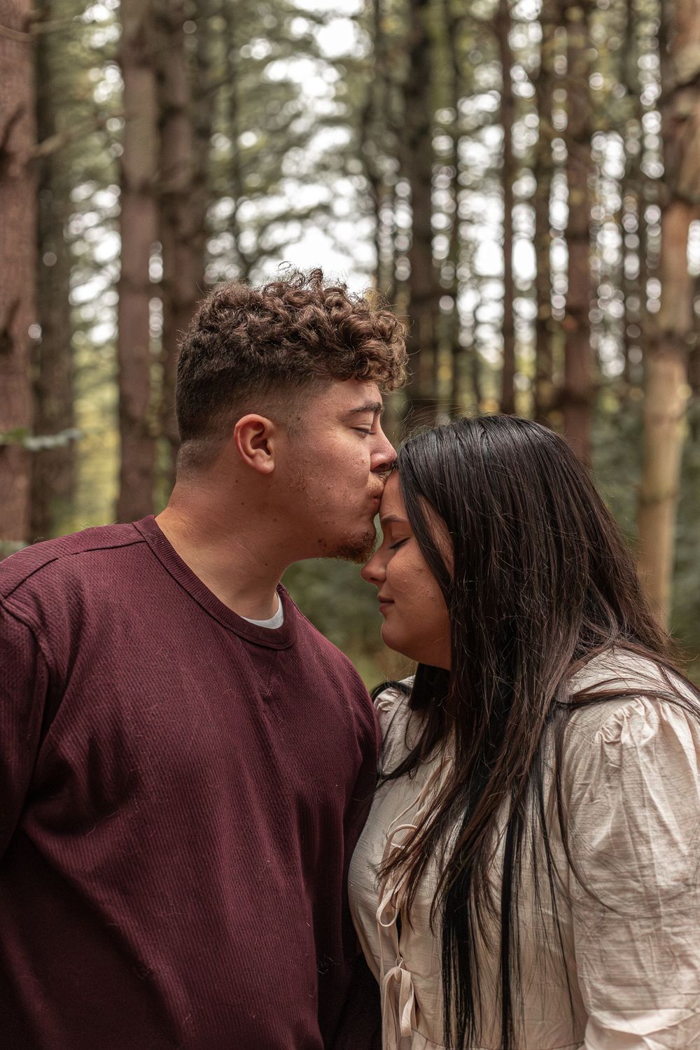 A romantic couple sharing an intimate moment in a forest setting with tall pine trees in the background.