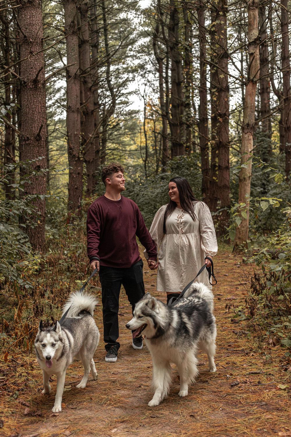 A couple walking their two Husky dogs on a forest trail surrounded by tall evergreen trees.